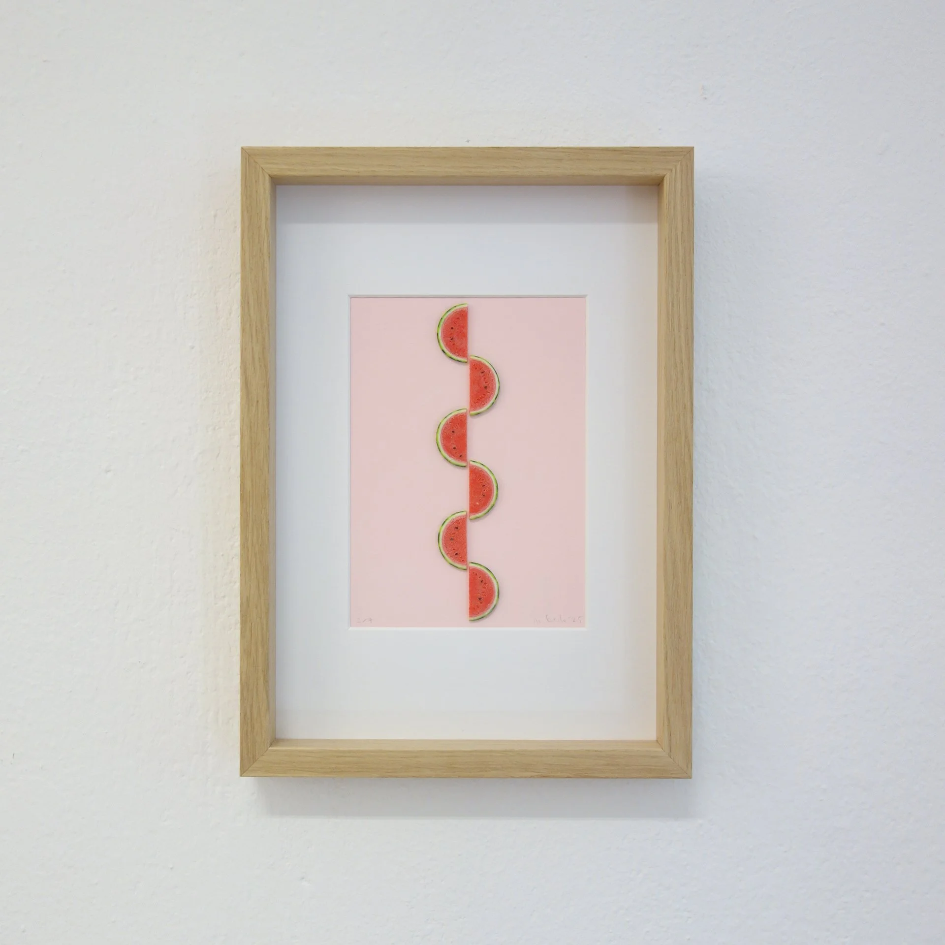 Fotografie eines gerahmten Kunstwerks mit mehreren Wassermelonenschnitten in einer vertikalen Linie auf rosa Hintergrund, an einer weißen Wand.