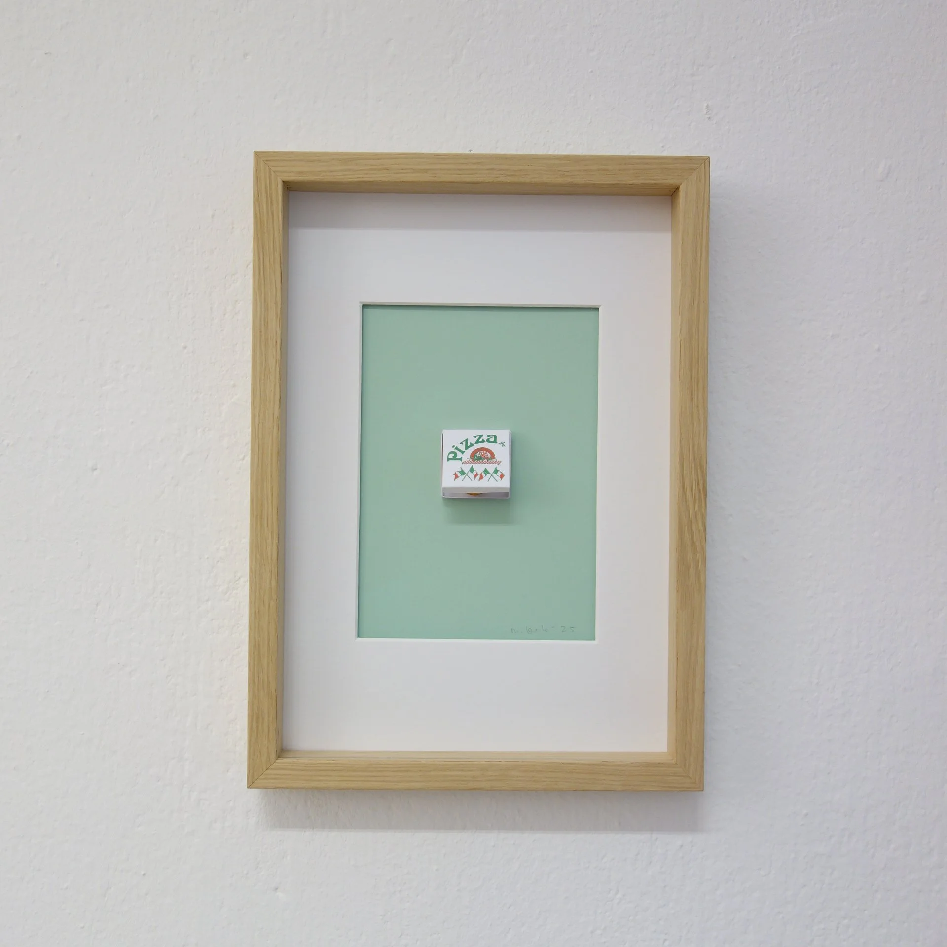 Ein Bilderrahmen aus hellem Holz an einer weißen Wand, darin ein grünes Blatt Papier mit einem kleinen Aufkleber, auf dem ein Pizza-Symbol und der Text "Pizza" zu sehen sind.