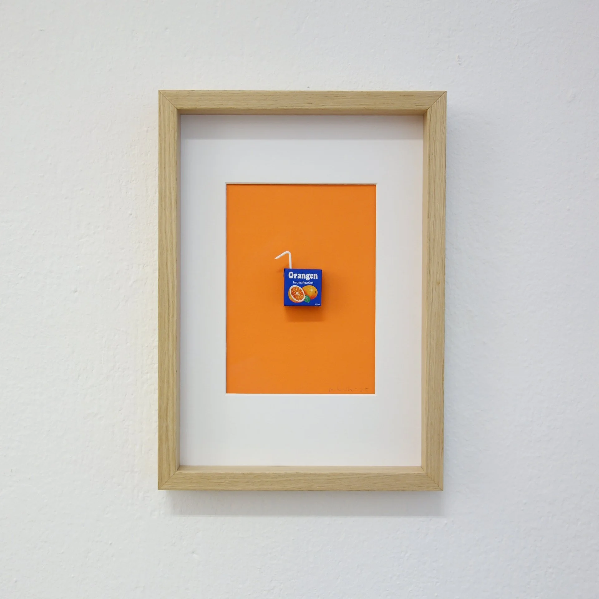 Ein Holzrahmen mit einem orangefarbenen Poster, das eine kleine blauen Packung Fruchtsaft zeigt, auf weißer Wand.