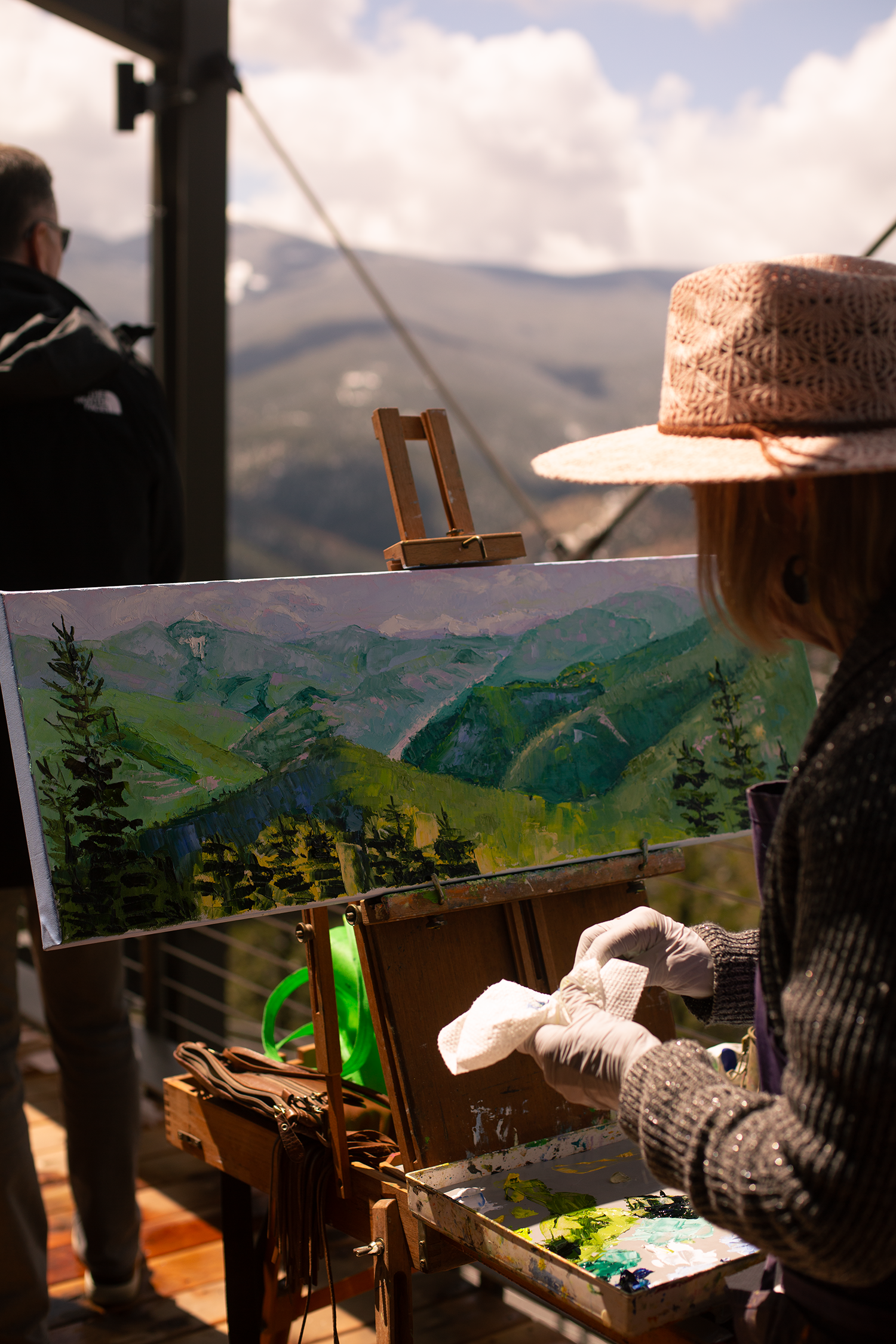 Painting a landscape on a canvas outdoors while another person watches nearby, with mountains and blue sky in the background near the Rocky Mountains, Colorado.