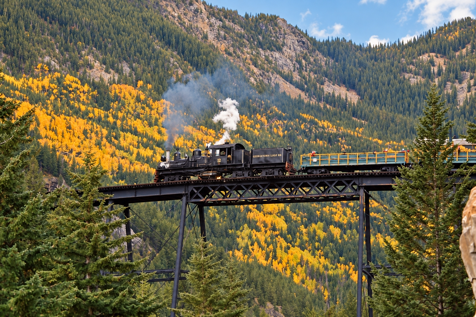 A black steam locomotive train traveling over a bridge in mountainous forested area with autumn-colored trees in the Rocky Mountains near Denver, Colorado.
