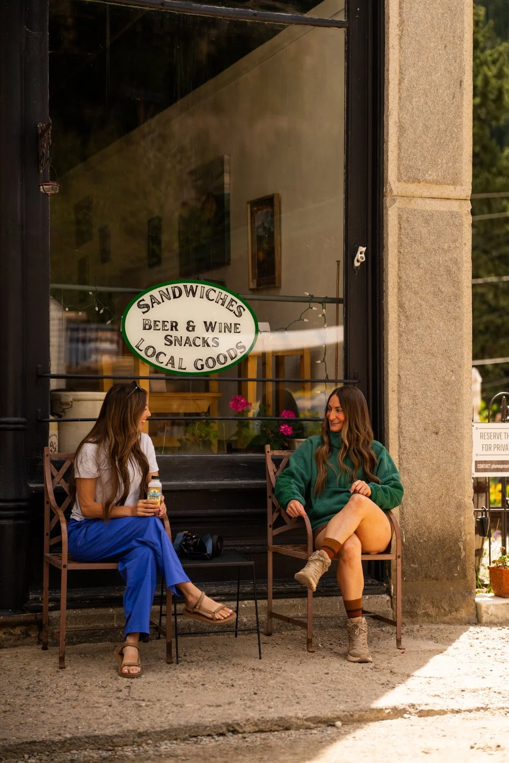Friends and family sitting outside a store with a sign that says sandwiches, beer & wine, snacks, local goods, smiling and talking to each other in the Rocky Mountains near Denver, Colorado.