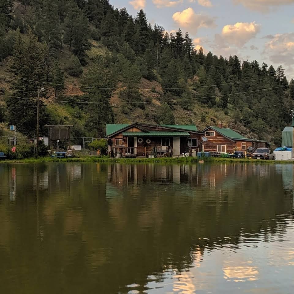 A serene lakeside scene with wooden cabins, surrounded by lush green trees and hills under a cloudy sky in the Rocky Mountains near Denver, CO.