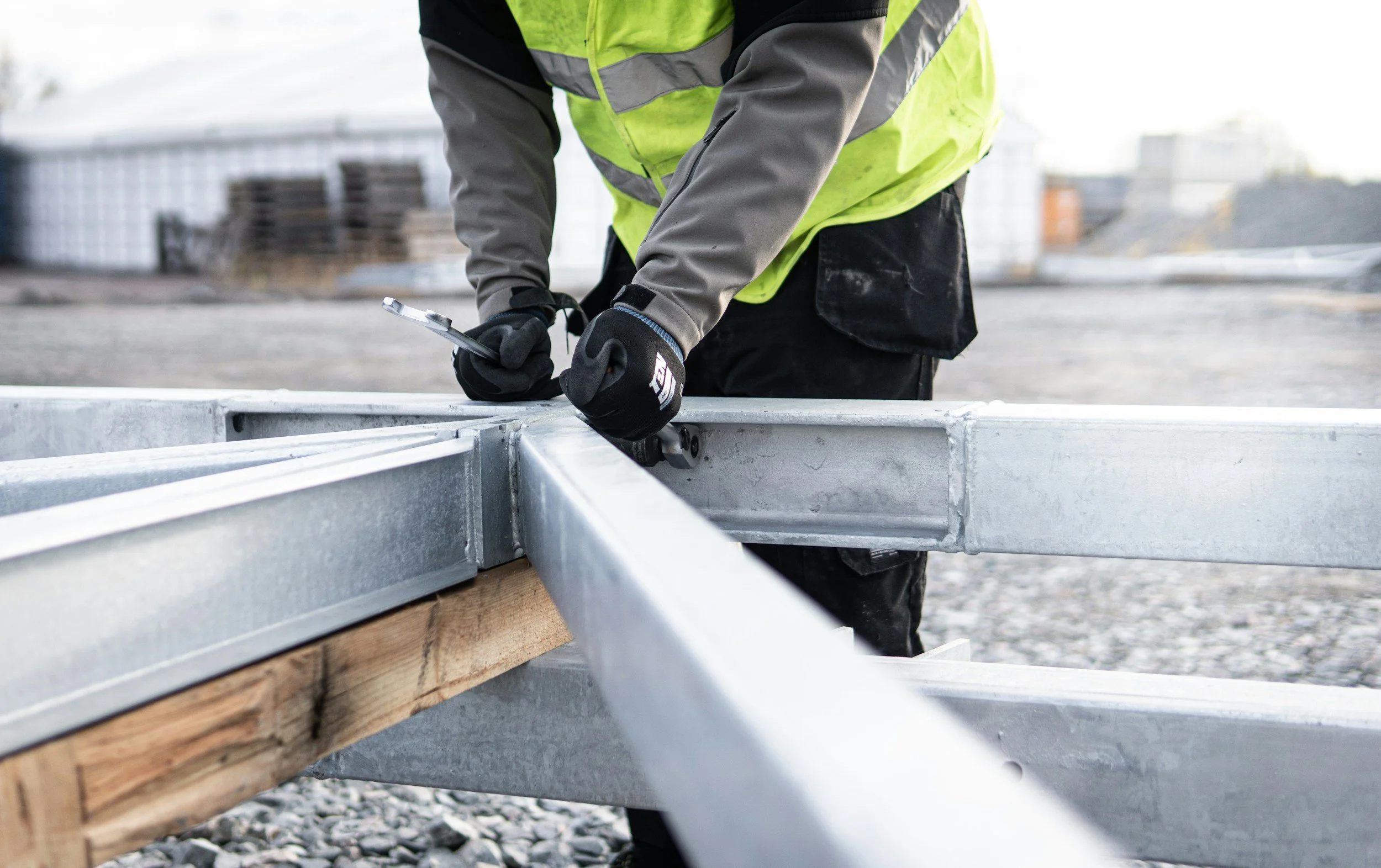 A construction worker wearing a high-visibility vest and gloves is securing a metal frame with a wrench on a construction site.