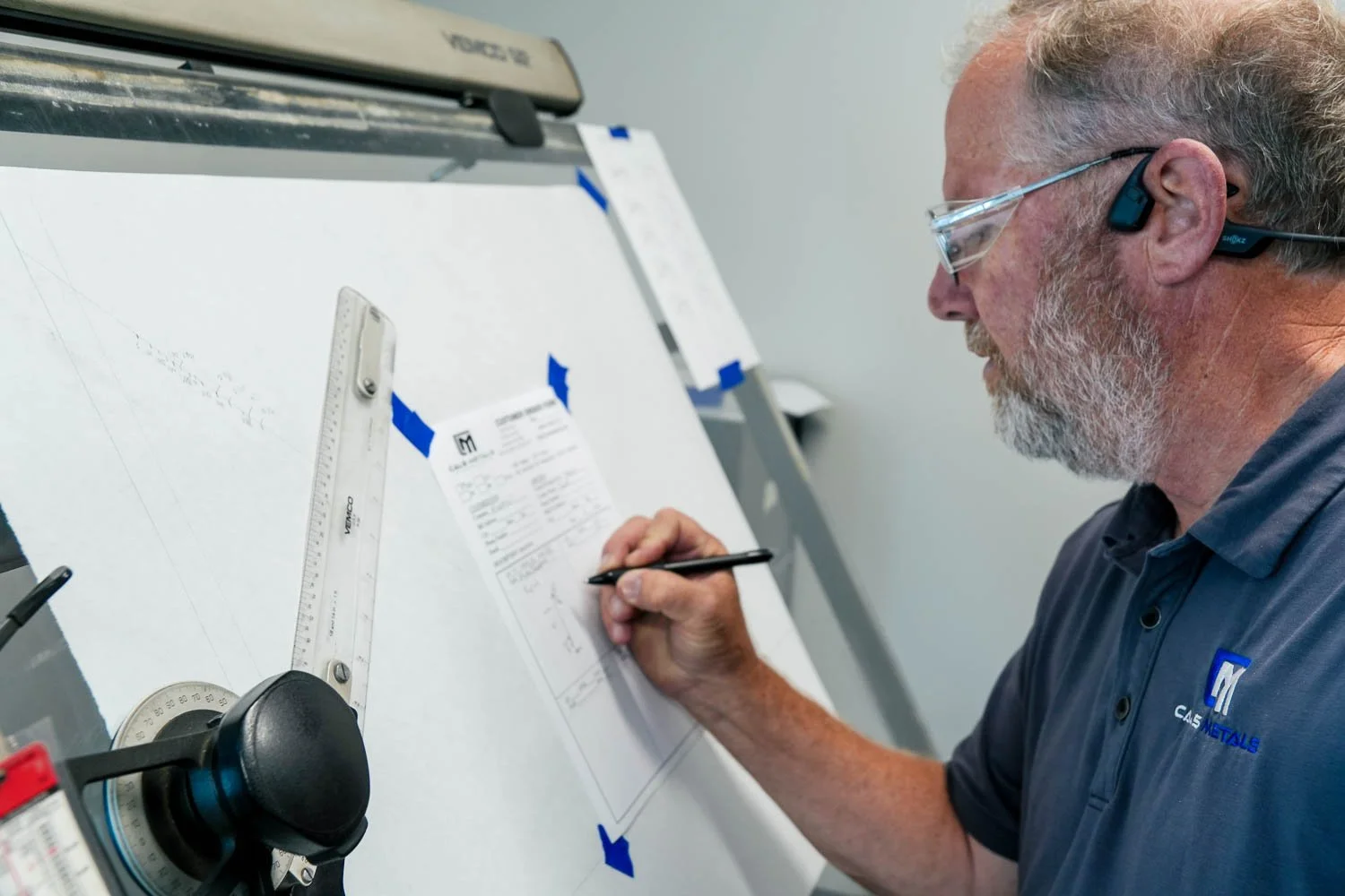 A man writing on a clipboard attached to a whiteboard with blue tape, wearing a gray shirt with the logo 'CAL'S METALS'.