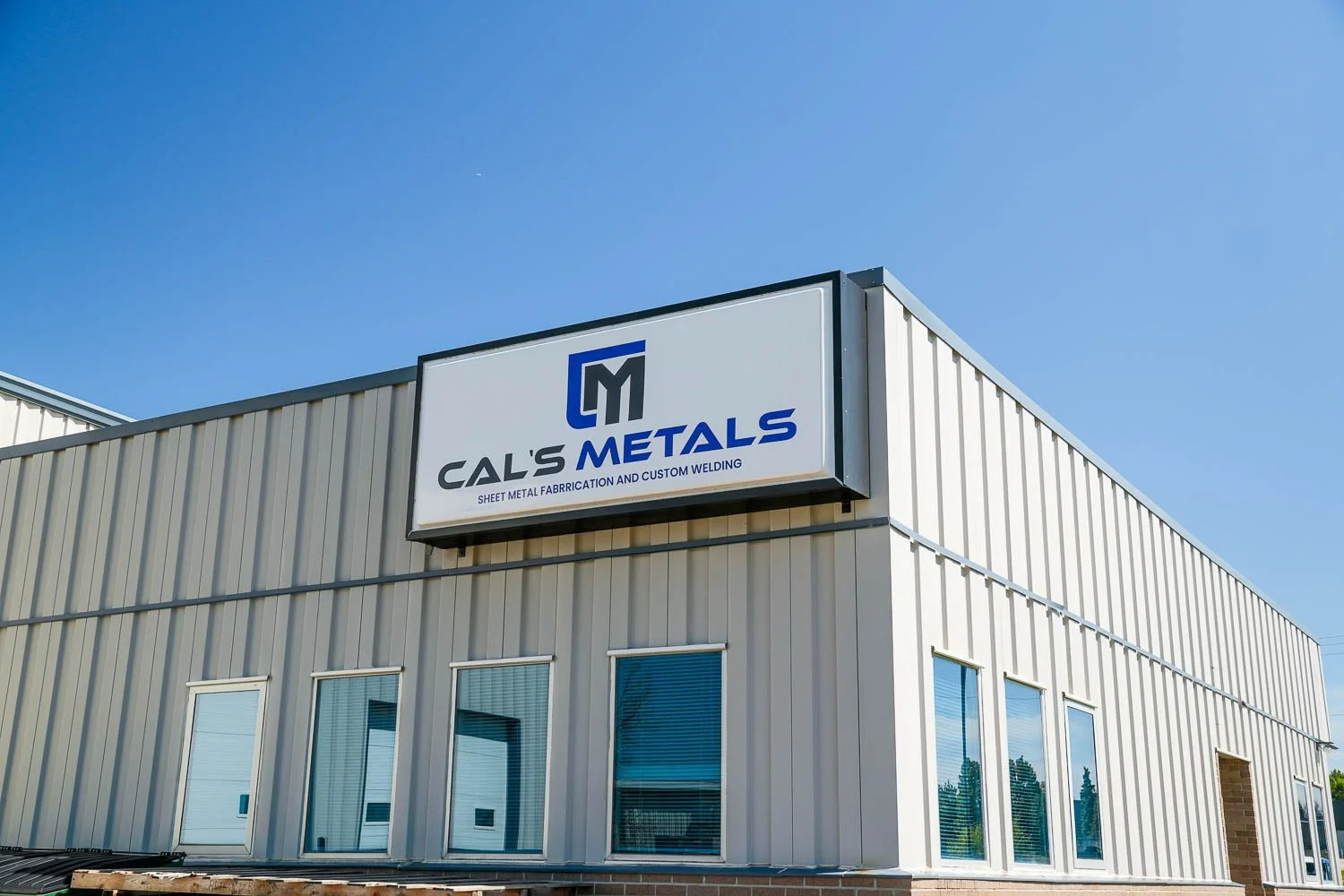 Exterior view of a metal building with a sign that says 'Cal's Metals' and 'Sheet Metal Fabrication and Custom Welding' under a clear blue sky.