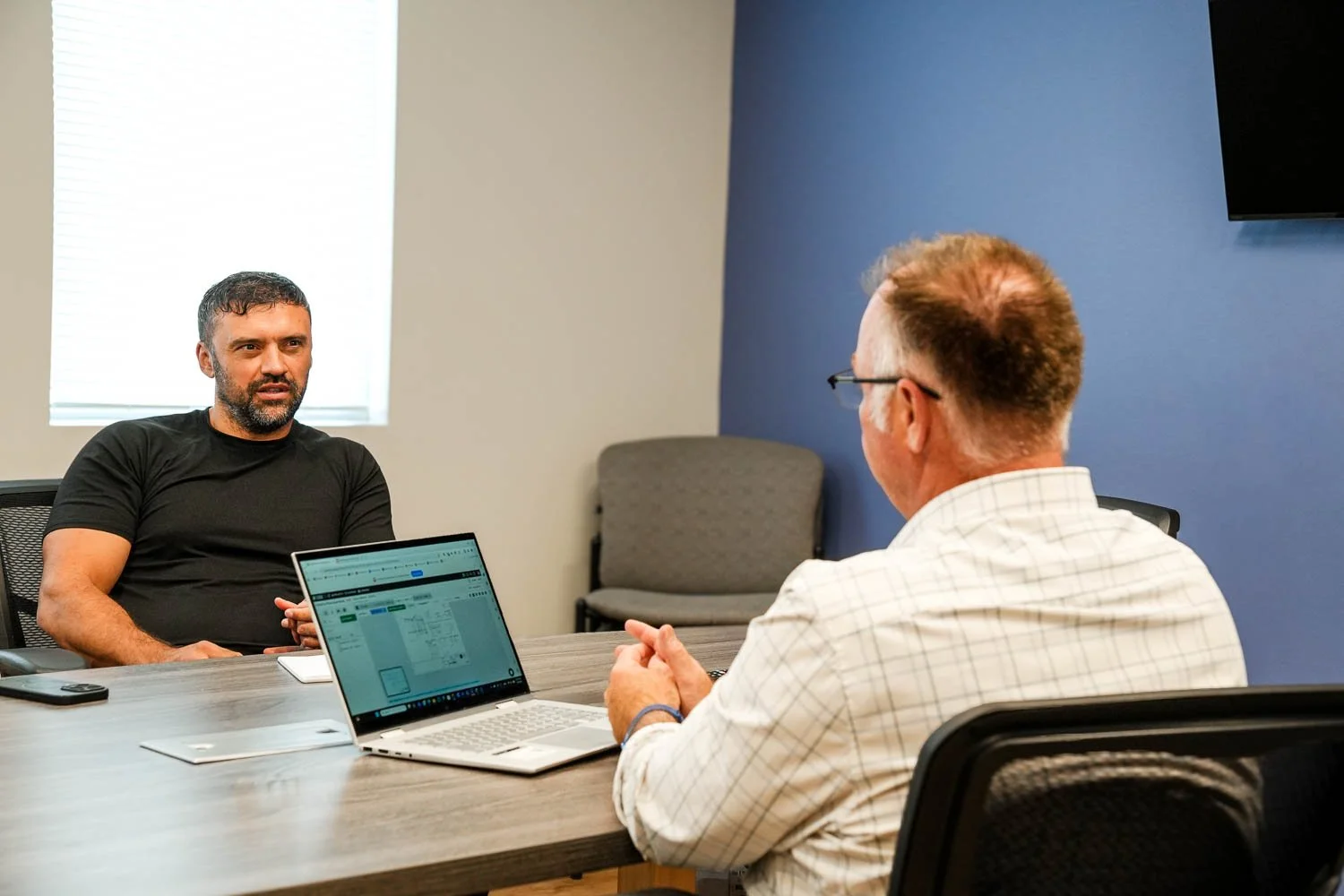Two men sitting across from each other at a conference table, engaged in a discussion. One holds a laptop displaying a calendar or schedule, the other is speaking. The room has a gray and blue wall with a window letting in natural light.