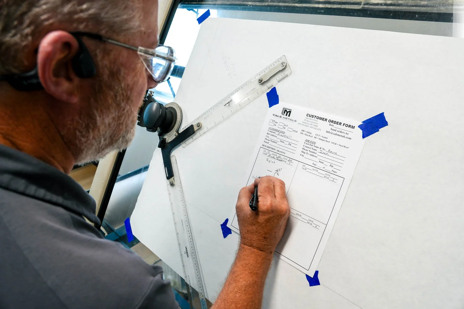A man is writing on a customer order form mounted on a white board, which is taped with blue painter's tape. He is using a pen and has a drafting T-square and a ruler on the board.