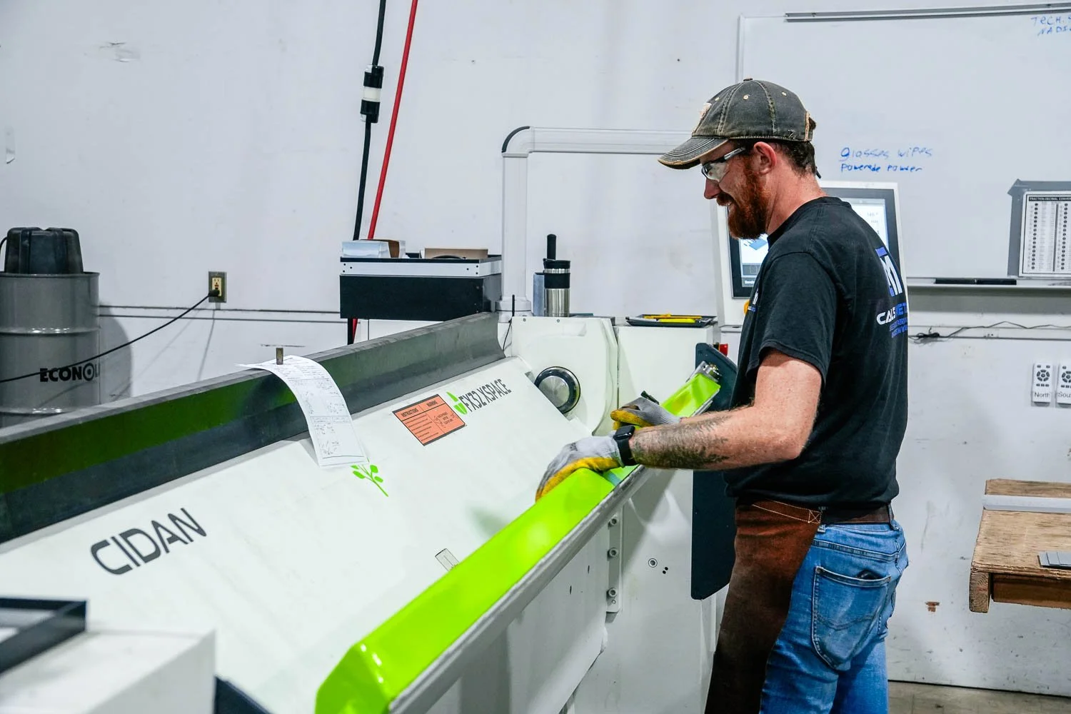 A man working in a metal fabrication shop, operating a large industrial cutting machine, wearing gloves, glasses, and a cap.
