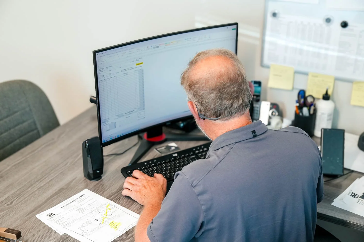 Man working at a desk with two computer monitors, one displaying a spreadsheet. The desk has papers, a phone, and office supplies, with sticky notes and a whiteboard in the background.