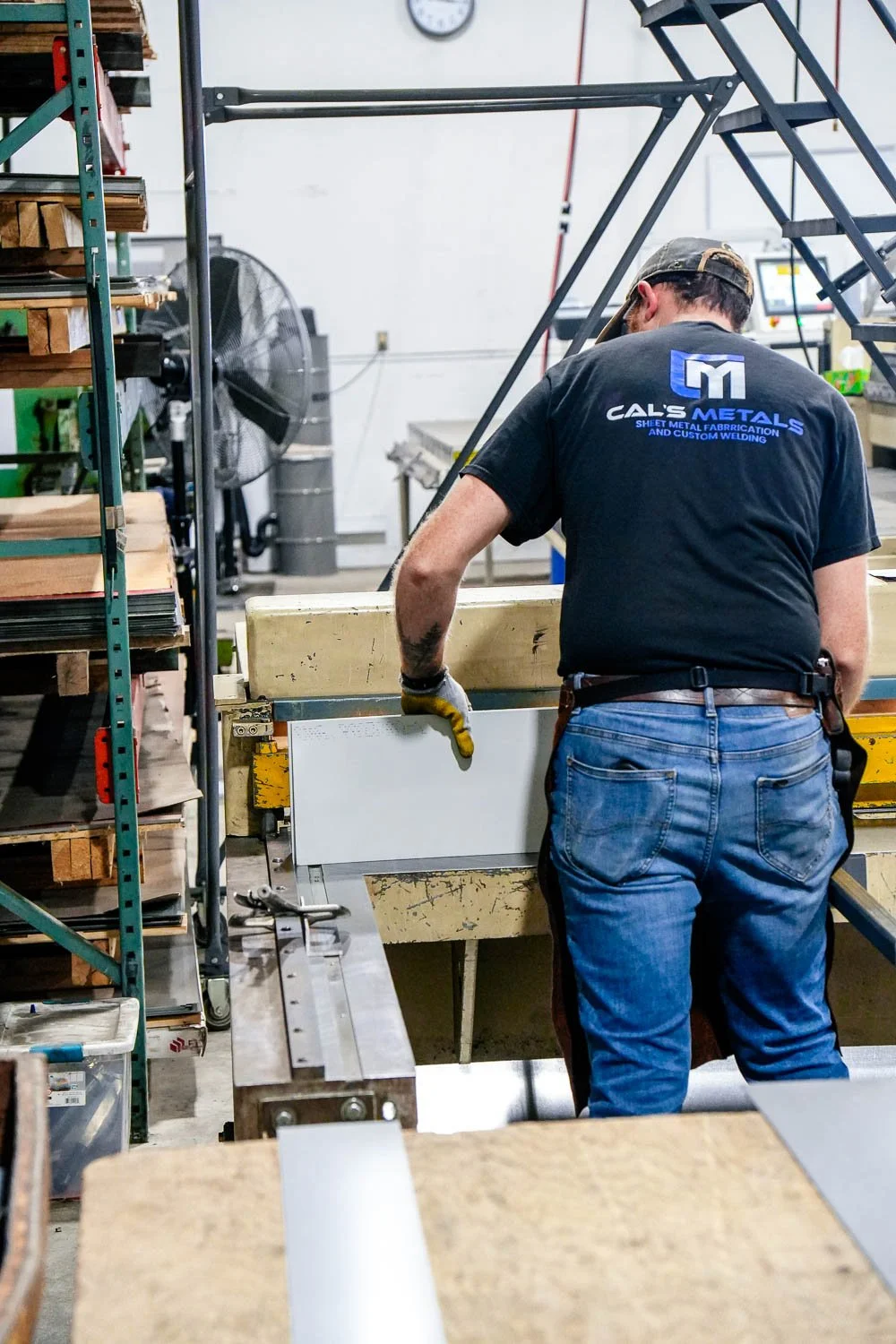 A man working in a metal fabrication shop, wearing a Cal's Metals T-shirt and using a saw on a white sheet of metal.