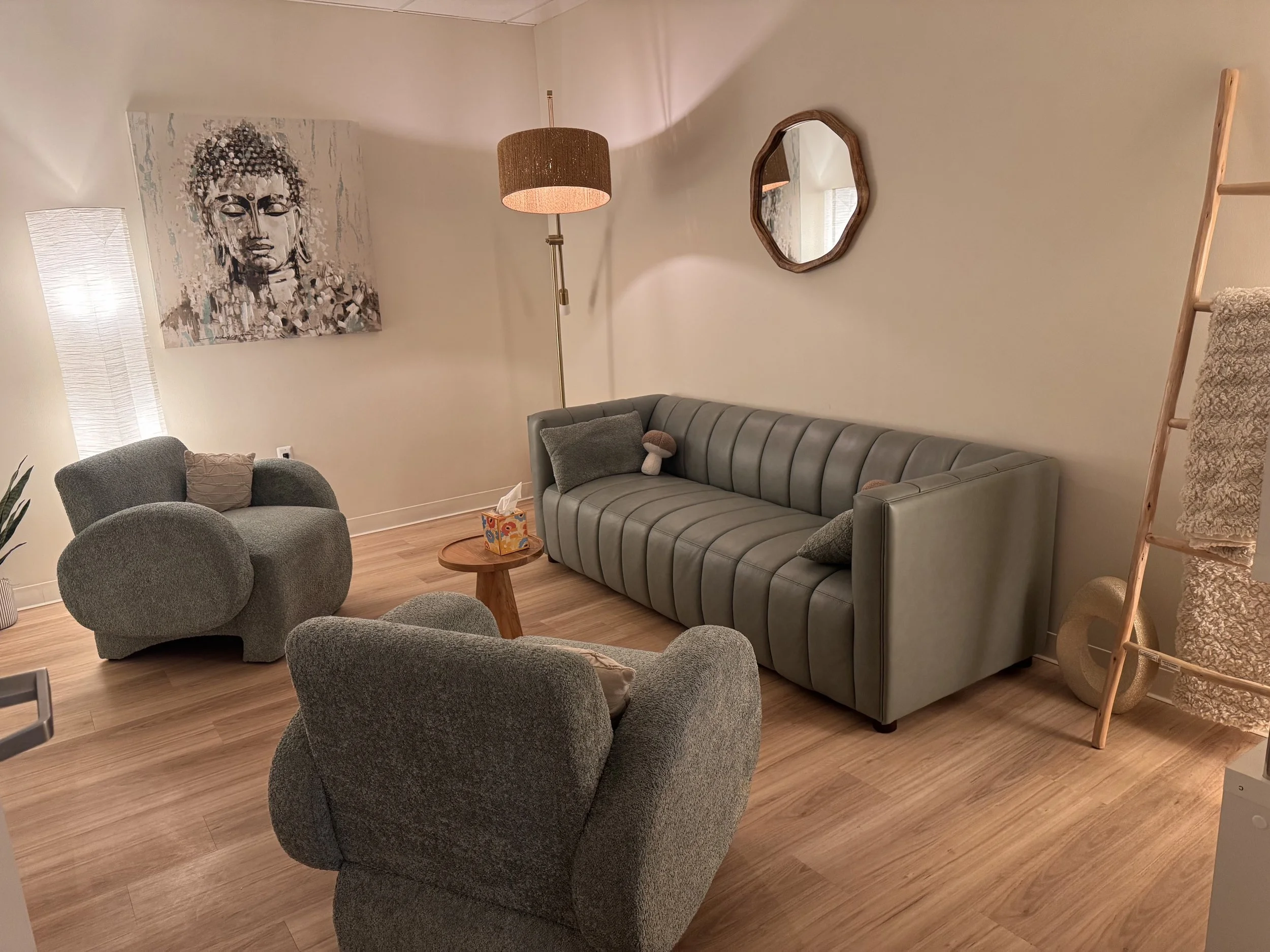Living room with gray upholstered armchairs, a gray sofa with pillows, a small wooden side table with a tissue box, a tall floor lamp with a brown shade, a rectangular abstract wall art depicting a face, a mirror, and a wooden ladder with blankets.