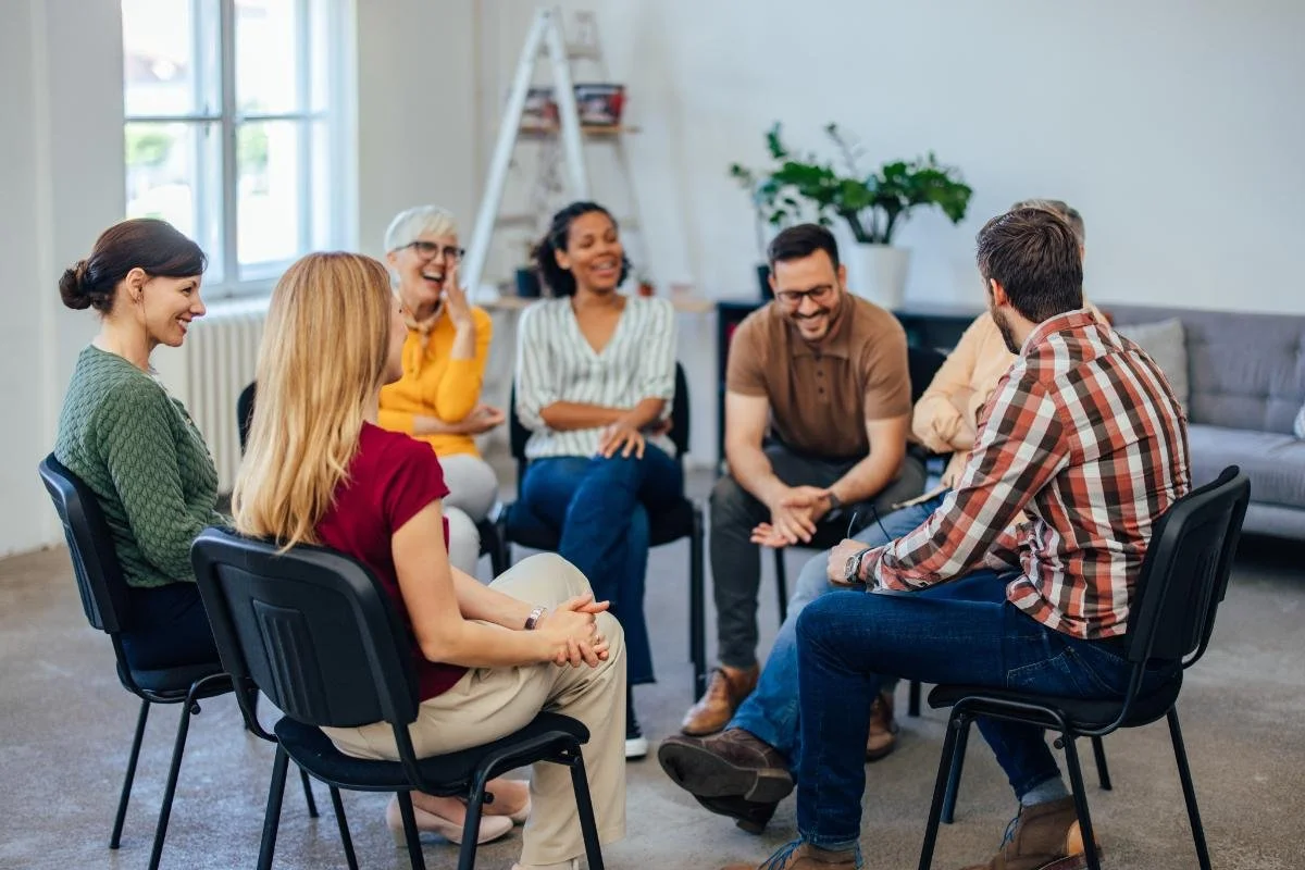 Group of people sitting in a circle and talking in a bright room with windows and a plant.