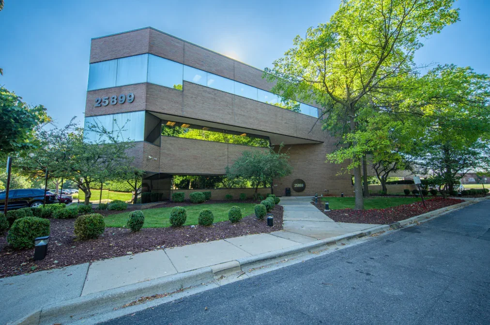 A modern office building with large reflective windows, brick exterior, and the address 25899 displayed on it, surrounded by landscaped greenery and trees.
