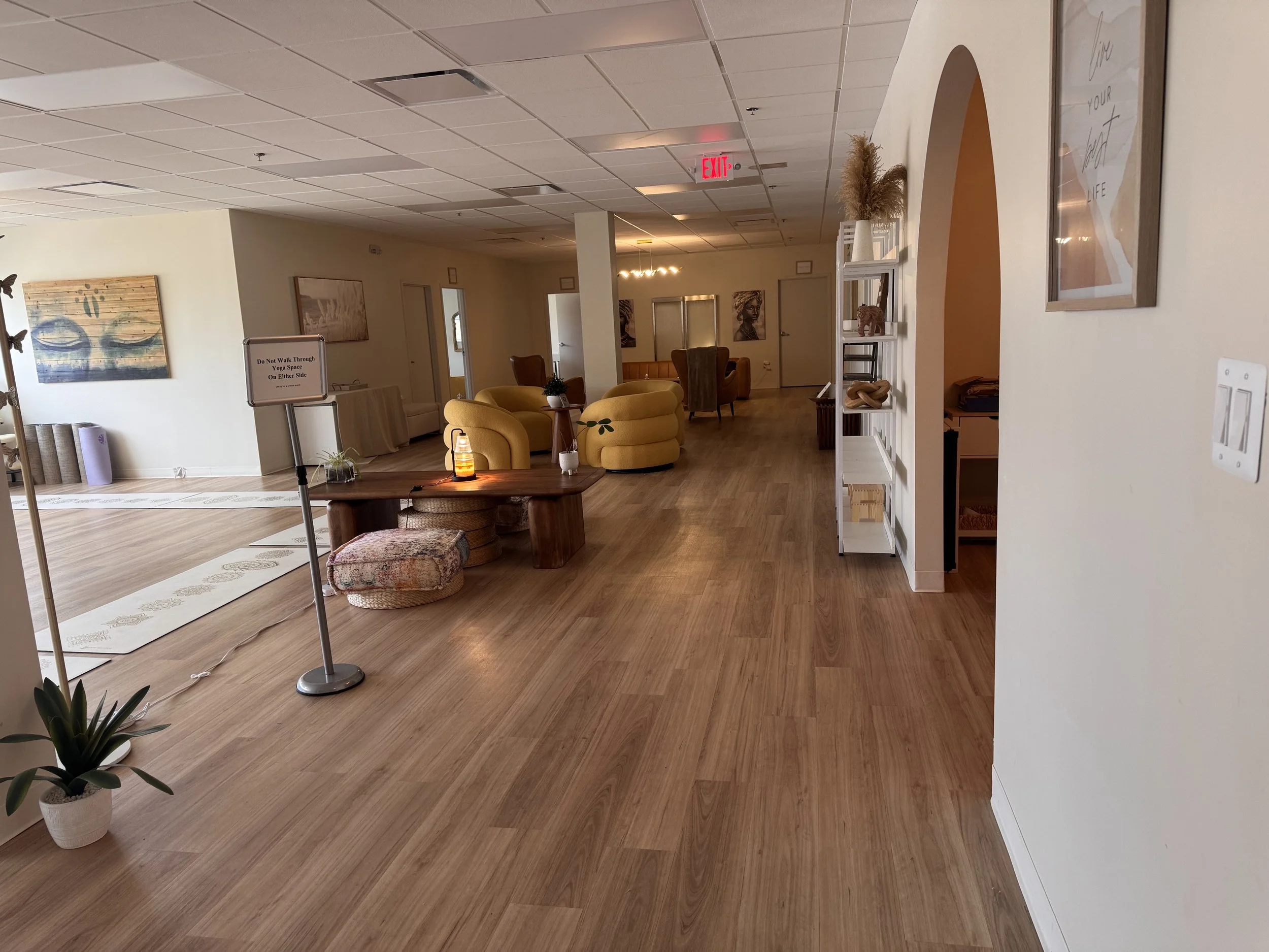 Lobby area with seating, including yellow armchairs and a small table with a lamp, potted plants, artwork on the walls, white shelving with decor, hardwood flooring, and a sign stating 'Do Not Walk Through Yoga Space on Either Side'.
