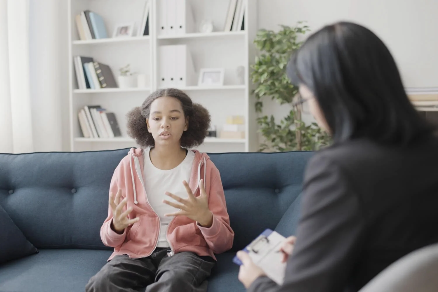 A young girl talking to a woman in a therapy session in a living room with a white bookshelf and a potted plant in the background.