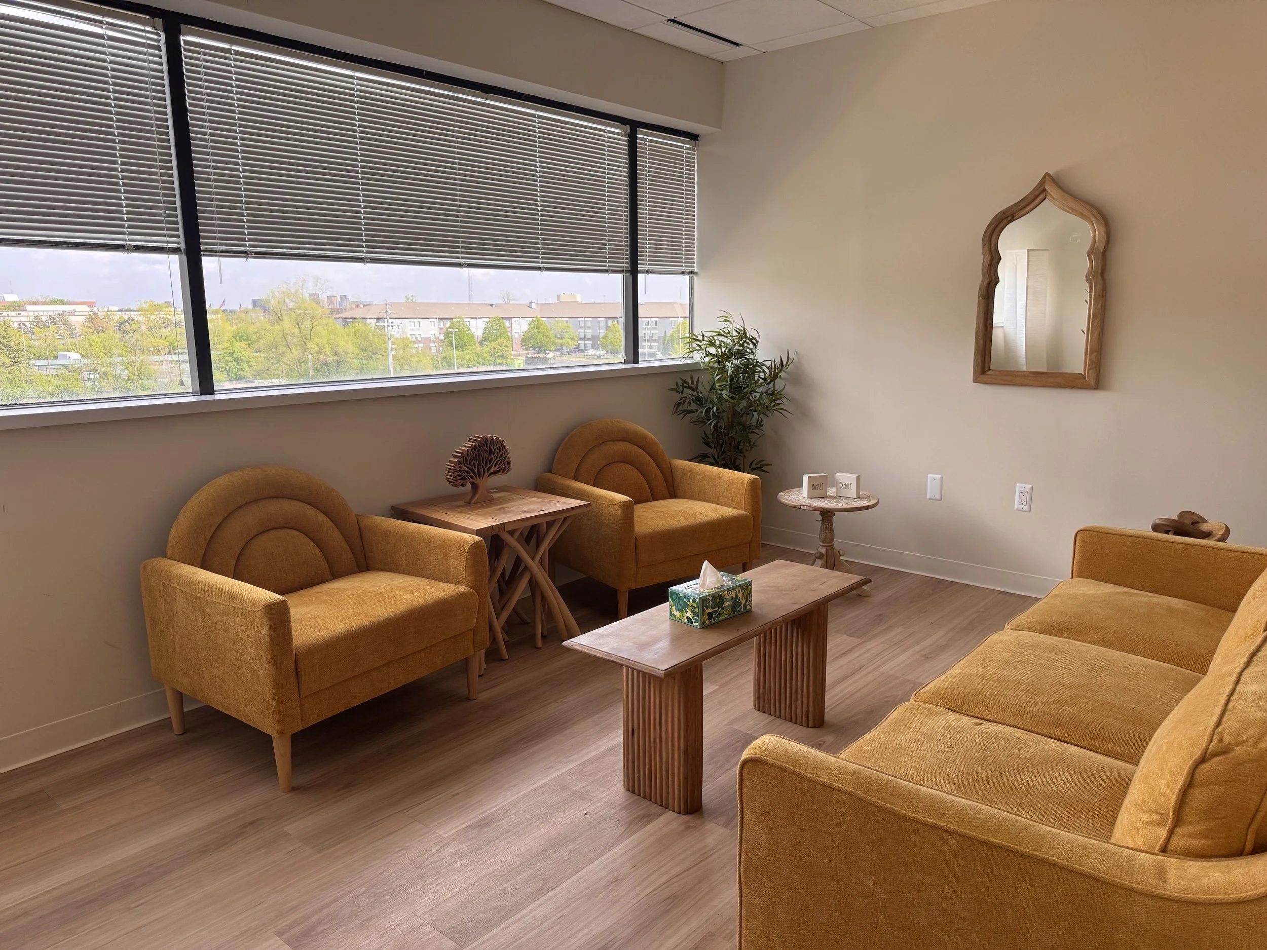 A cozy waiting or lounge room with mustard/yellow upholstered seating, small tables, a wooden table with a decorative item, a potted plant, the wall mirror, and a large window with blinds.