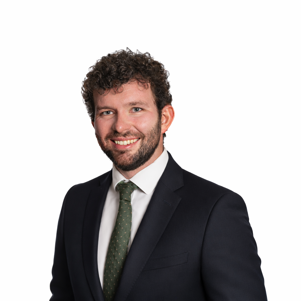 A smiling man with curly brown hair, a beard, wearing a black suit with a white shirt and a green tie, against a white background.