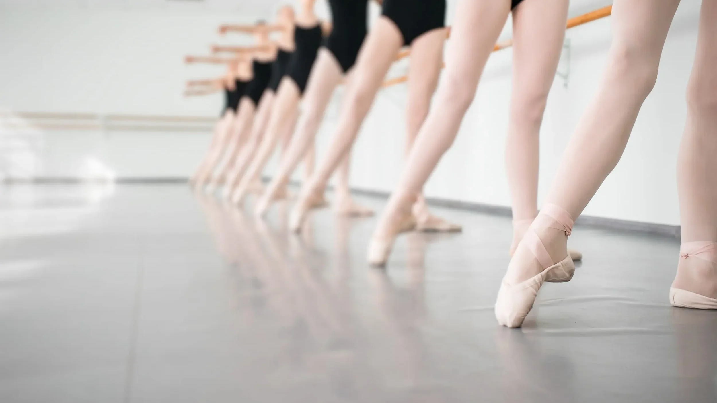 Ballet dancers in black leotards and pink tutus on pointe, stretching at a ballet barre in a dance studio.