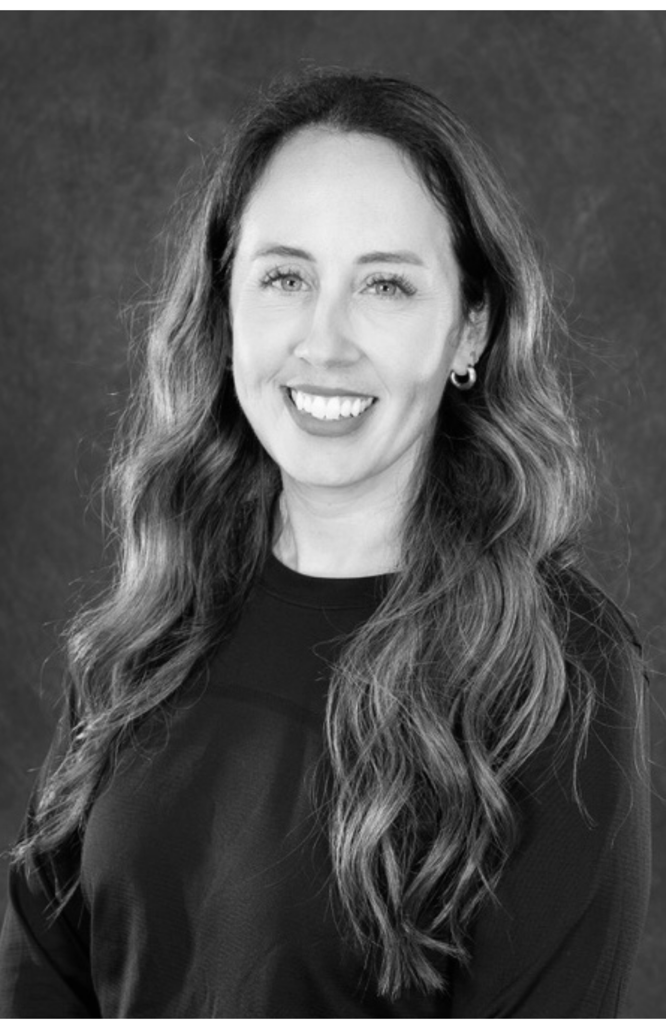 Black and white portrait of a woman with wavy hair, wearing earrings and a dark shirt, smiling and looking at the camera.