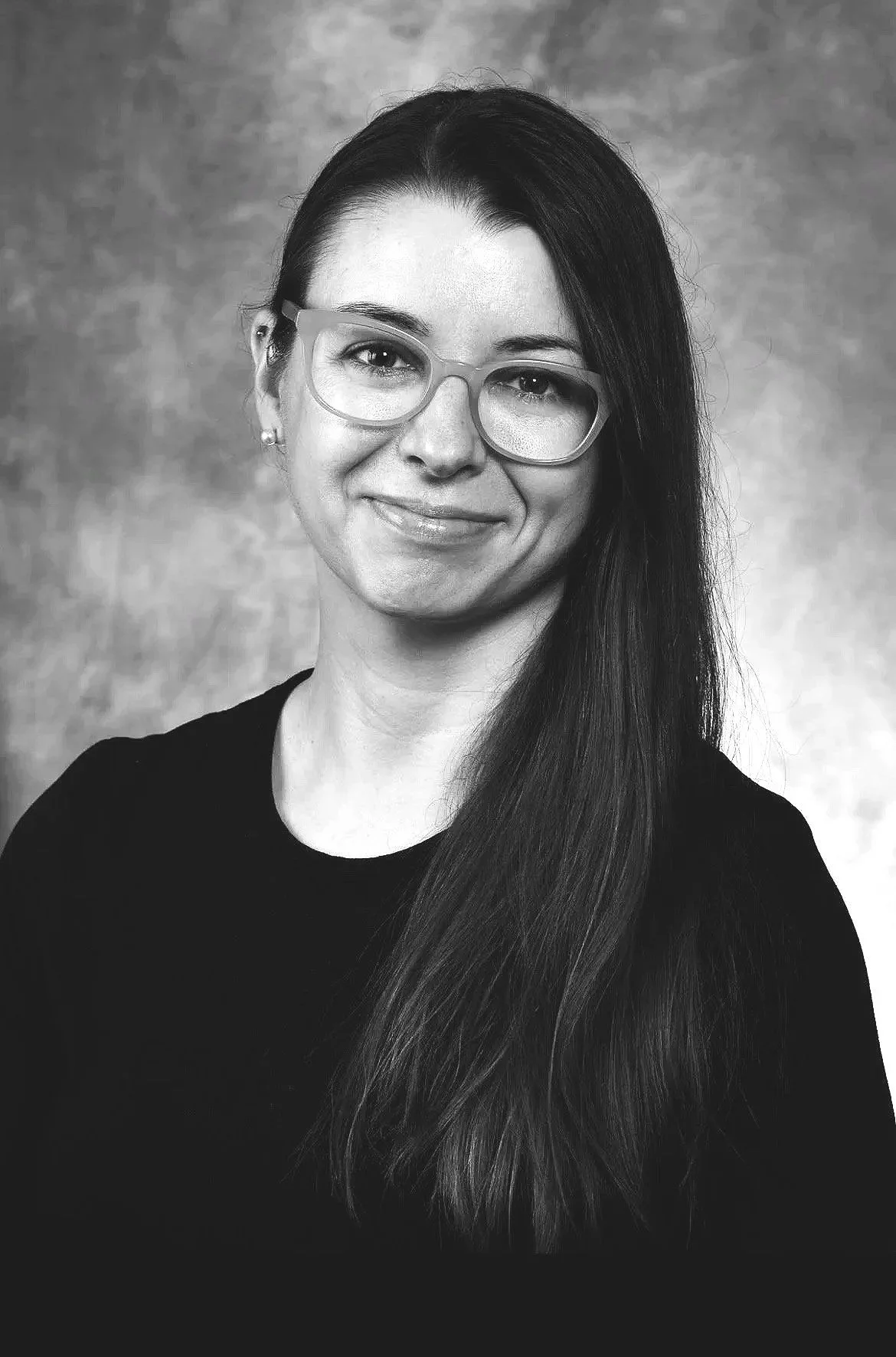Black and white portrait of a smiling woman with long dark hair, wearing glasses and earrings, dressed in a black top, against a textured background.