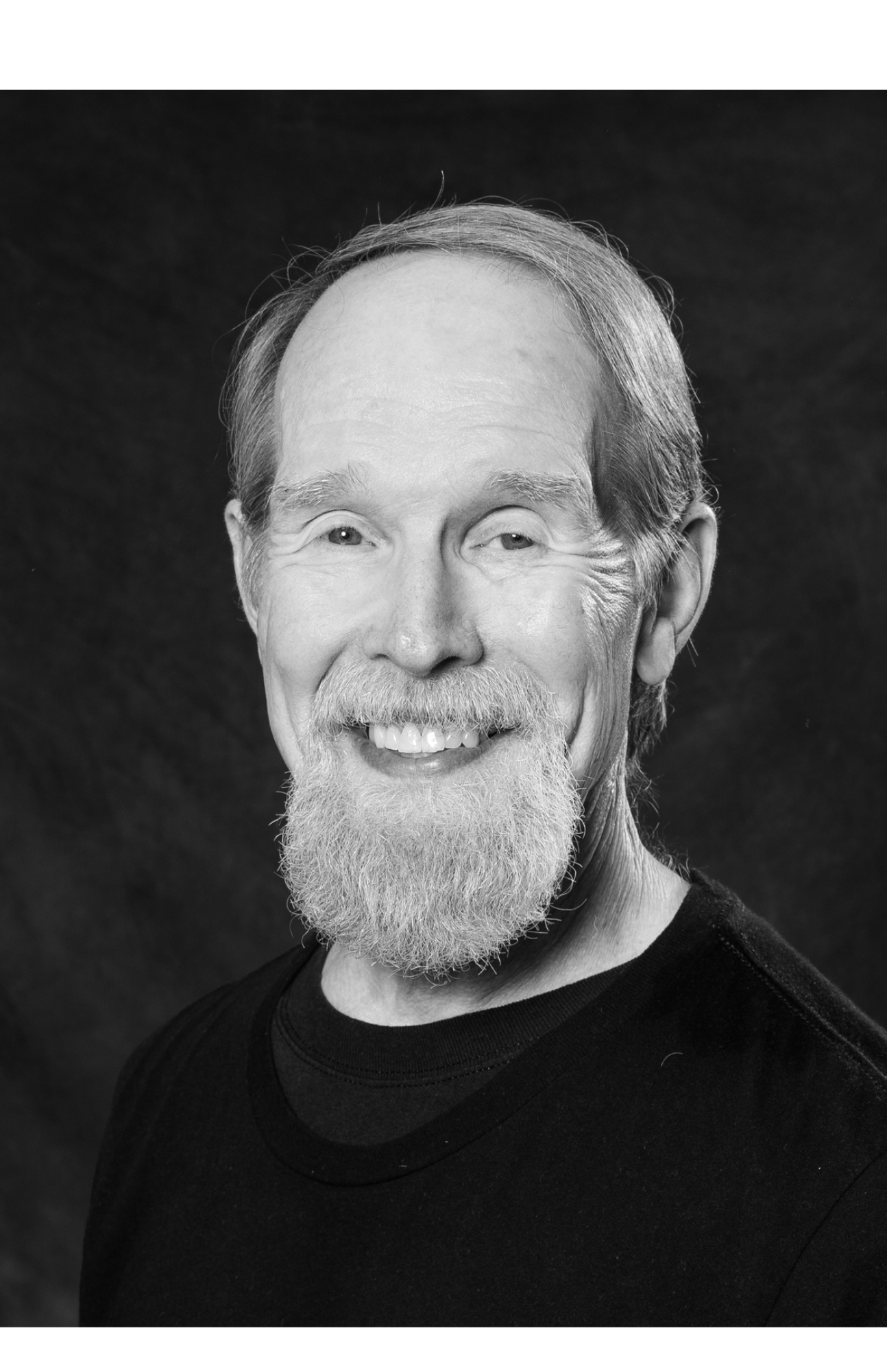 Black and white portrait of a smiling elderly man with a beard and mustache, wearing a dark shirt against a dark background.