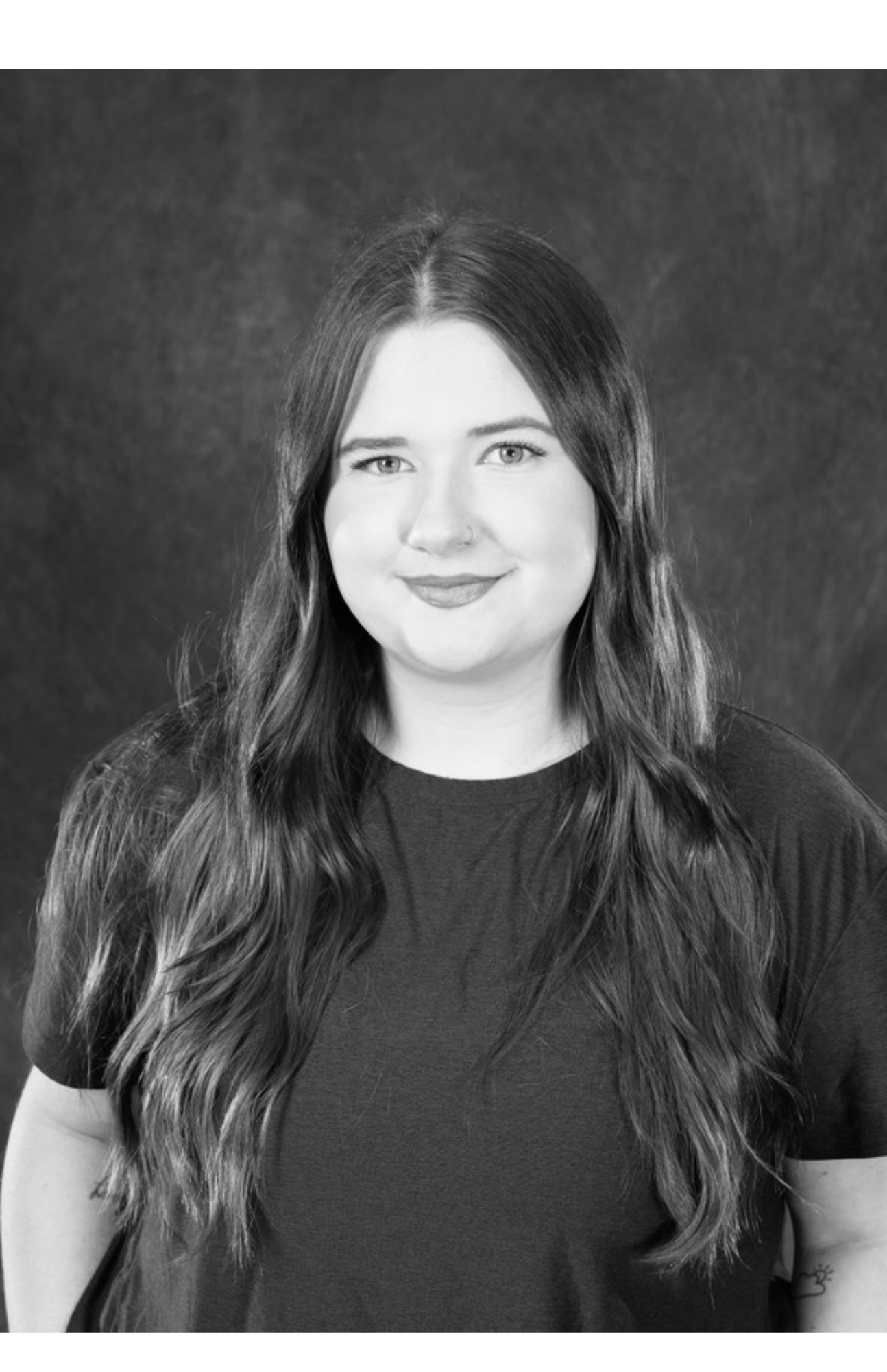 Black and white portrait of a woman with long wavy hair, wearing a t-shirt, smiling slightly, with a dark background.