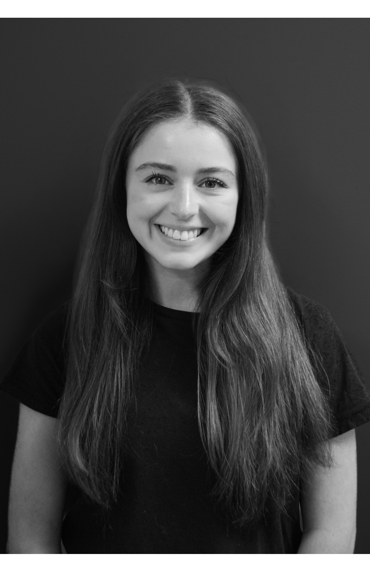 Black and white portrait of a young woman with long hair, smiling, wearing a dark shirt, against a plain background.