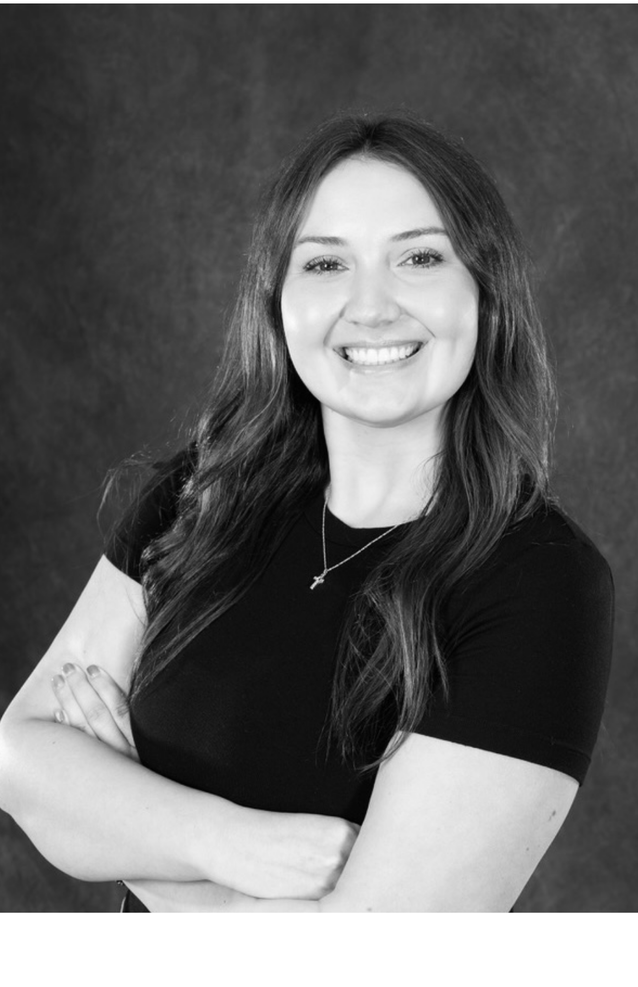 Black and white portrait of a woman with long wavy hair, smiling, arms crossed, wearing a black top and a necklace, against a dark background.
