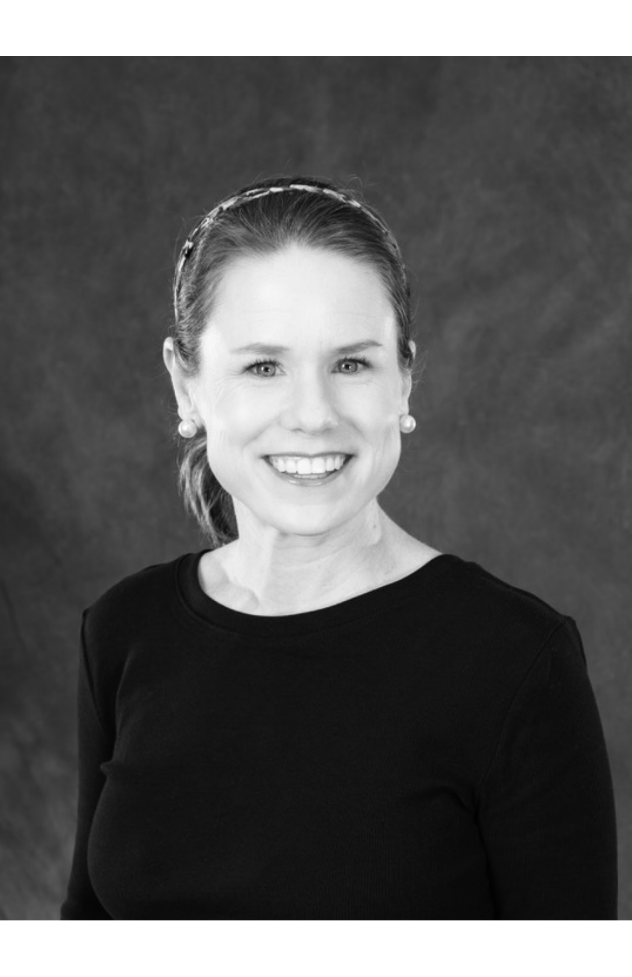 Black and white portrait of a smiling woman with pearl earrings, wearing a black top, looking directly at the camera, with a dark textured background.