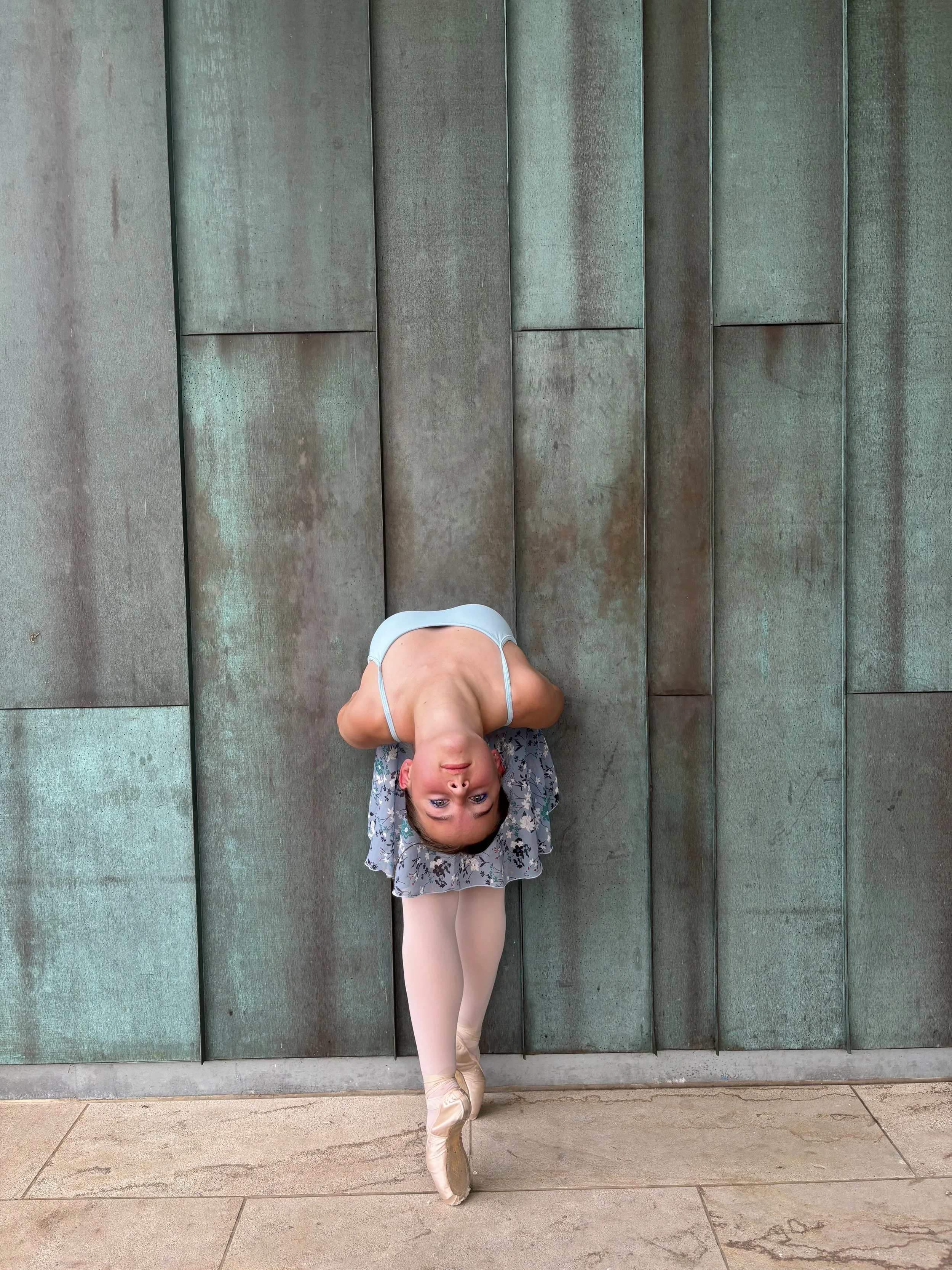 A ballet dancer in a tutu and pointe shoes practicing ballet on a sidewalk against a green metal wall, with her head upside down and feet pointed.
