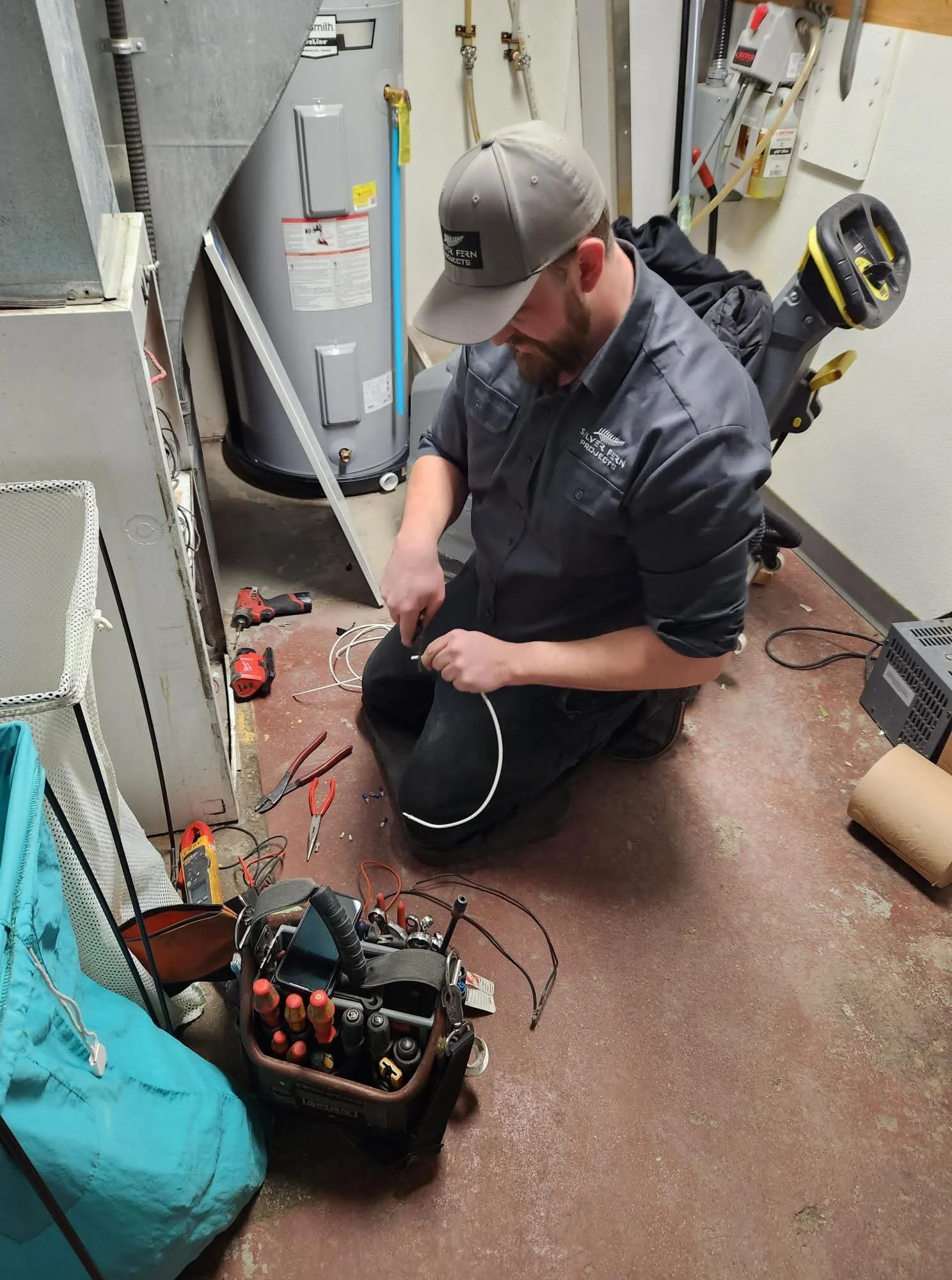 A man kneeling on the floor working on an electrical project, with tools and wiring scattered around him in a utility room with water heater and equipment on the wall.