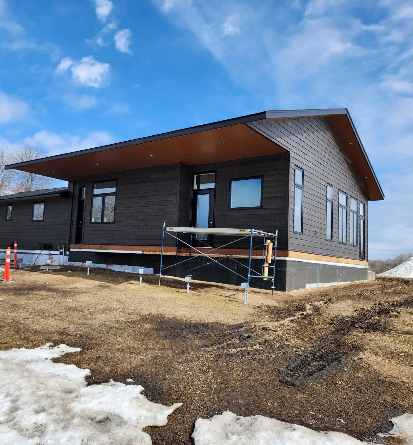 Modern black house under construction on a clear day with blue sky and some clouds, surrounded by dirt and patches of snow.