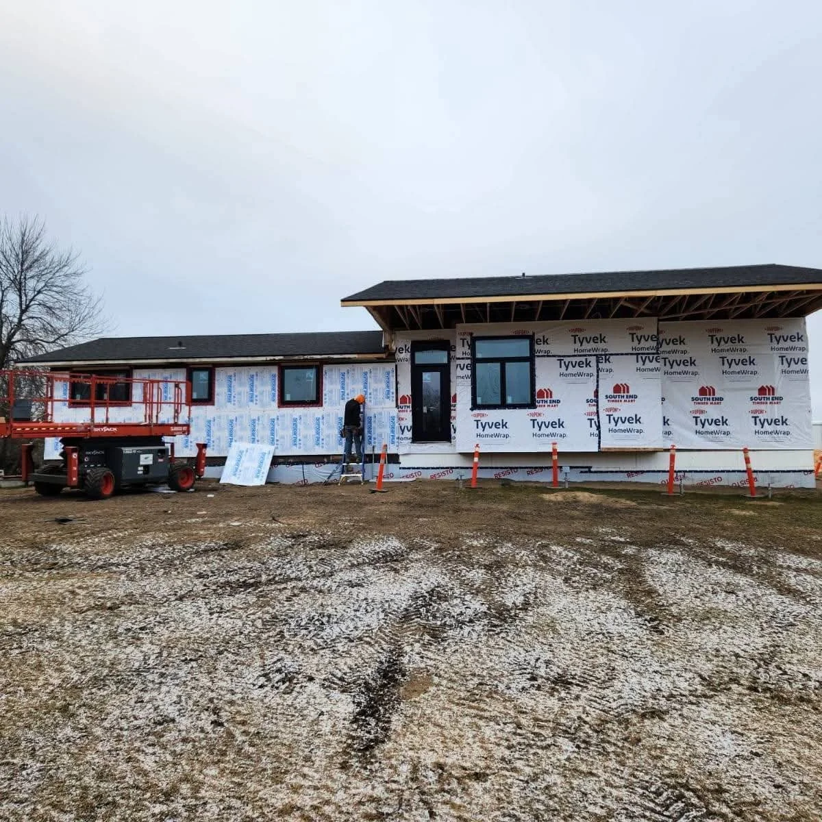 A house under construction with Tyvek HomeWrap exterior sheathing, a worker on a ladder, and construction equipment on a dirt lot.