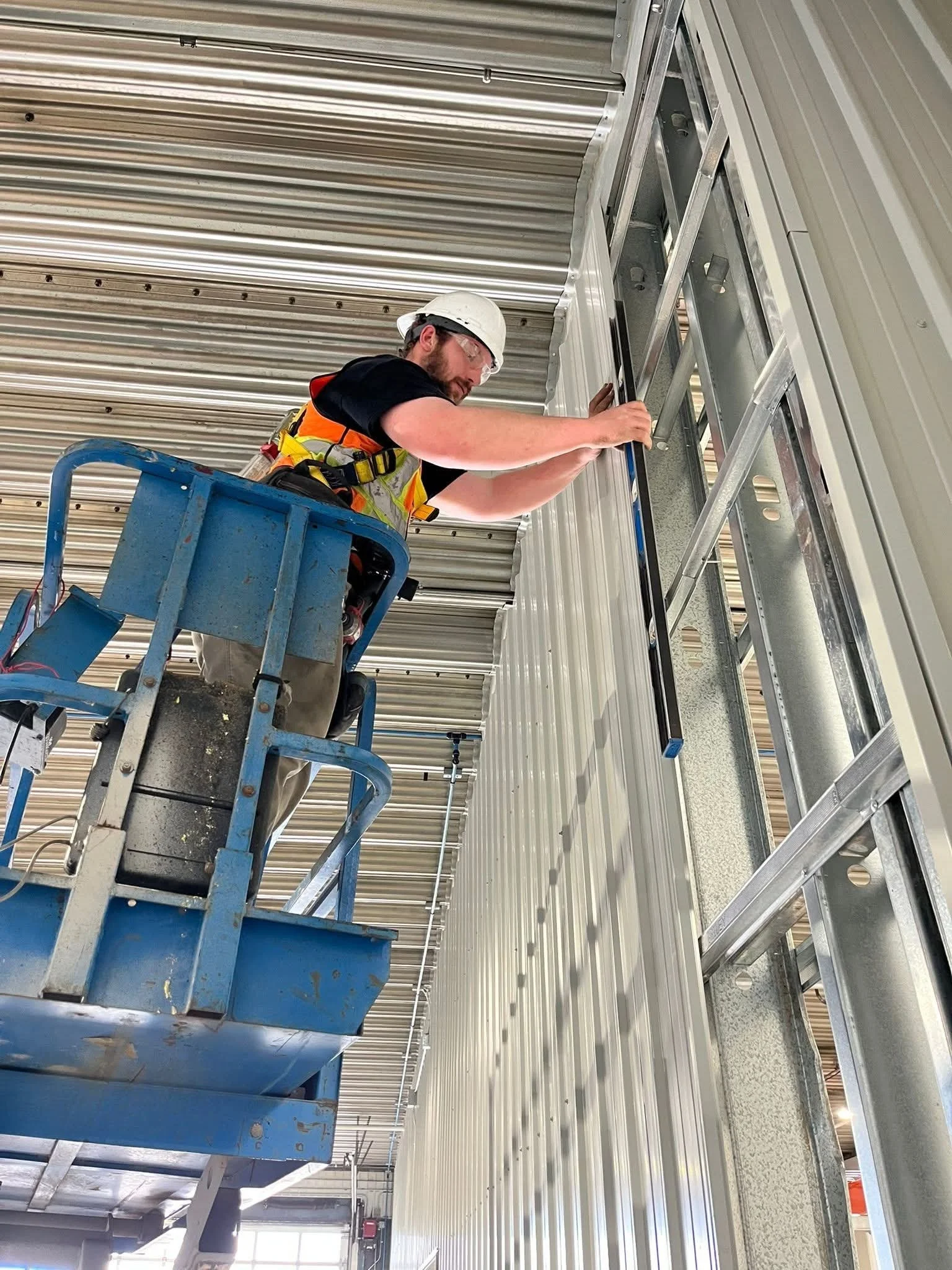 A worker is on a blue lift installing or repairing a metal sign or panel on a building's exterior wall. The worker is wearing safety glasses, a white hard hat, and a high-visibility safety vest, working under a corrugated metal roof.