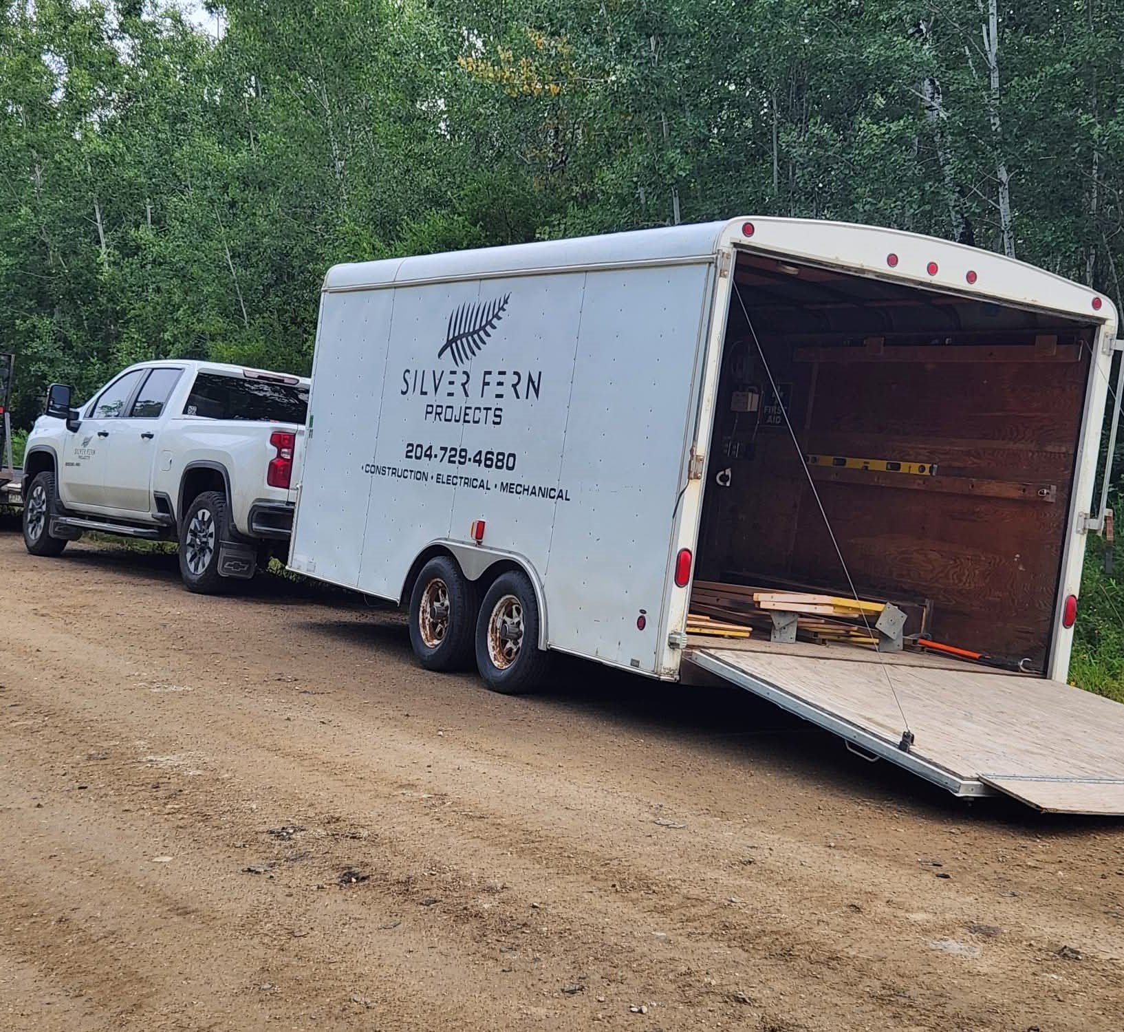 A white pickup truck parked beside an enclosed trailer with its rear ramp open, situated on a dirt area with trees in the background.