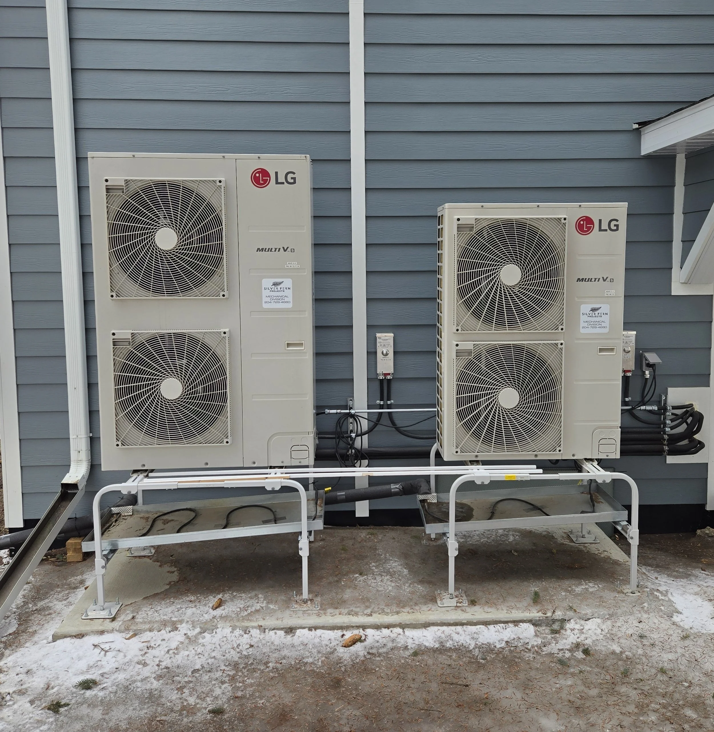 Two LG multi-v air conditioning units mounted on a wall with blue horizontal vinyl siding. The units are on white metal brackets and are connected by black wires and pipes. The ground beneath the units shows dirt, patches of snow, and some small pine cones.