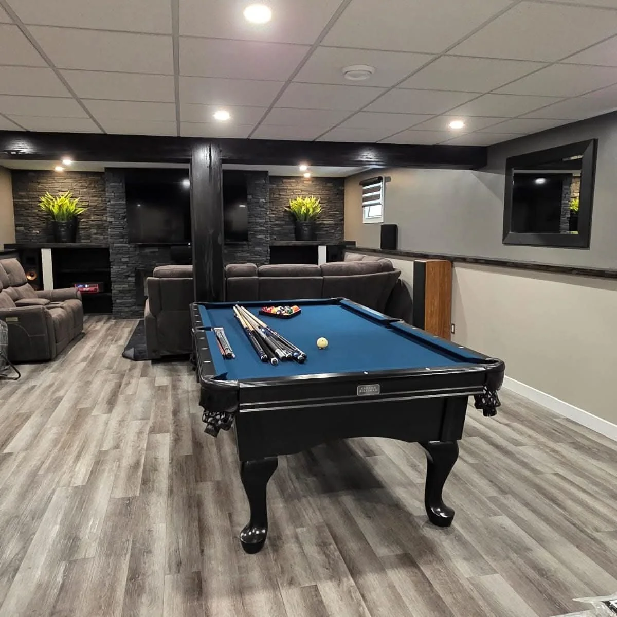 A basement recreation room with a pool table in the center, dark wood flooring, a large wall-mounted television, plush seating, a mirror, and decorative plants.