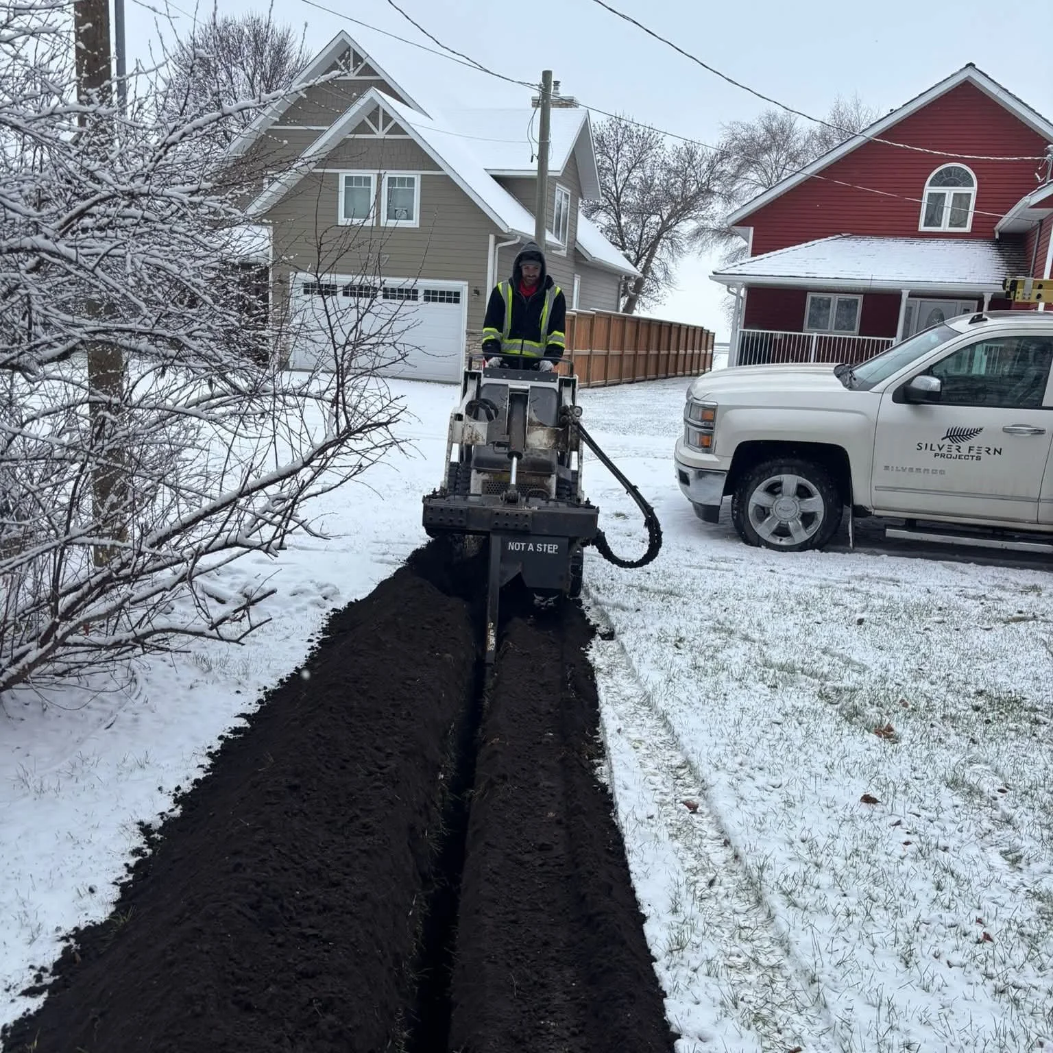 A worker in a reflective vest operating a trenching machine that is digging a long trench in a snow-covered yard near houses, with a white truck parked nearby.