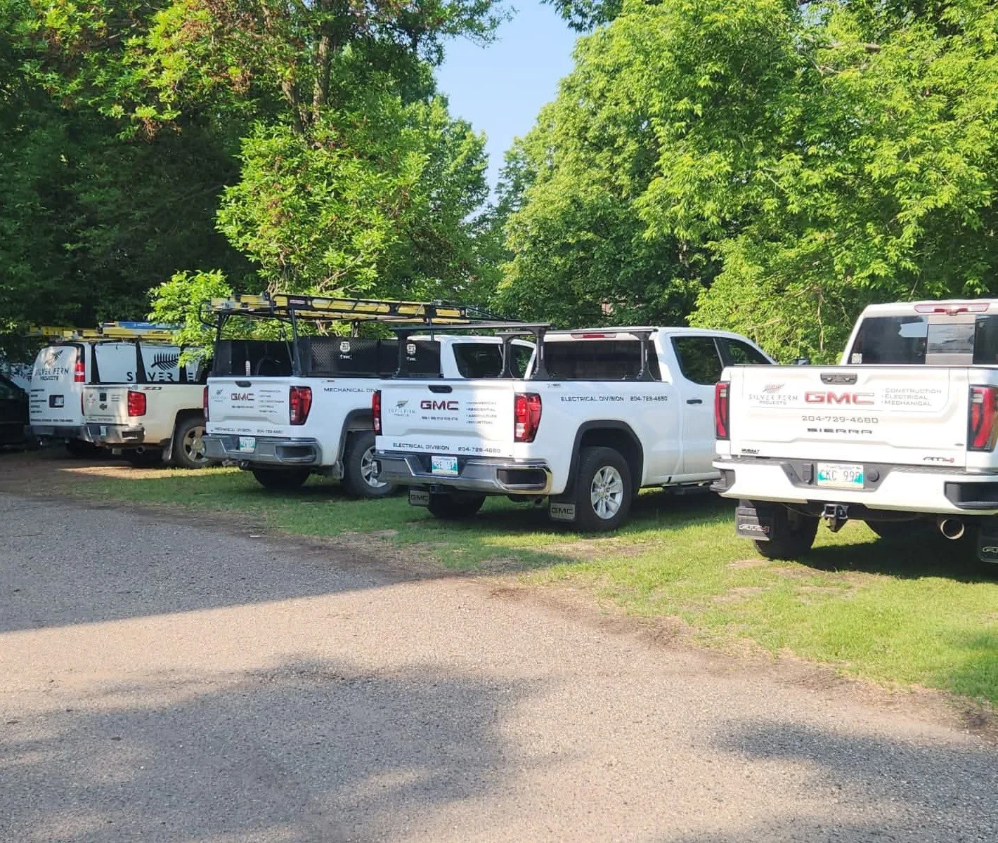A row of white GMC service trucks parked on a grassy area under green trees.
