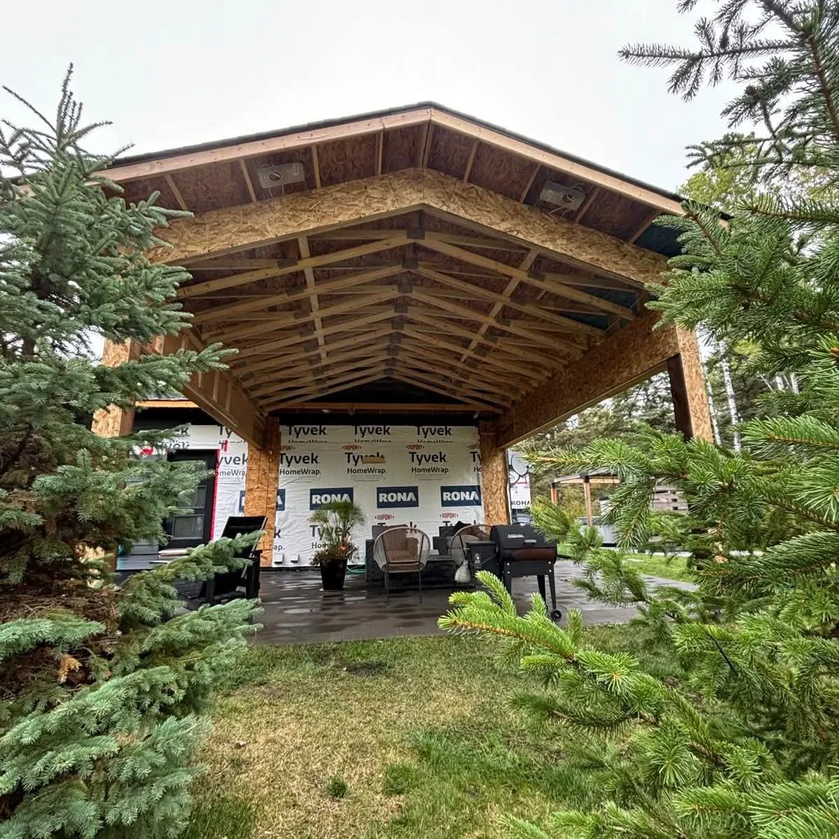 Construction of a covered outdoor patio or deck with exposed wooden framing, surrounded by evergreen trees.