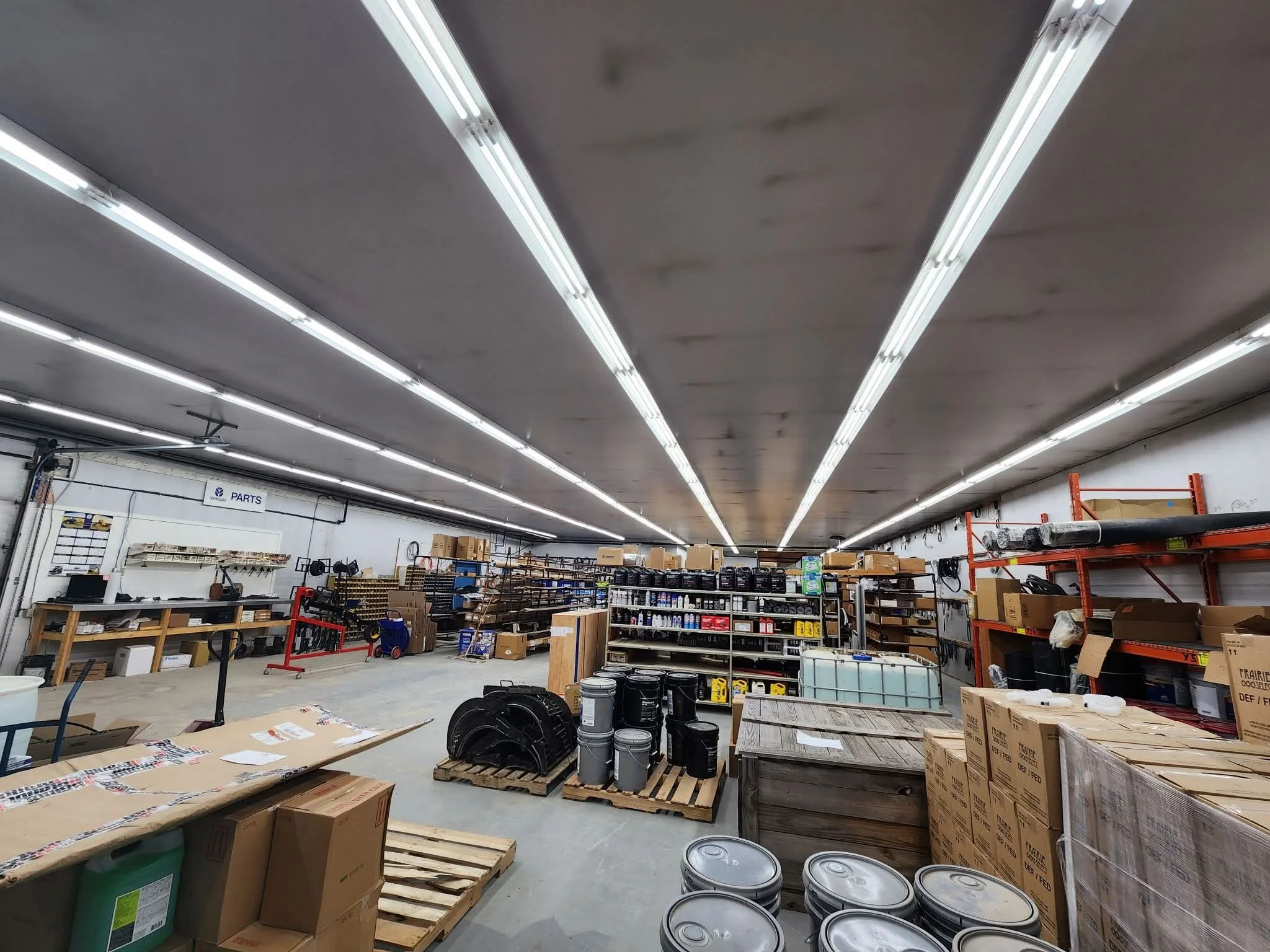 Interior of a warehouse or workshop with shelves containing boxes, cans, and tools, and fluorescent lighting on the ceiling.