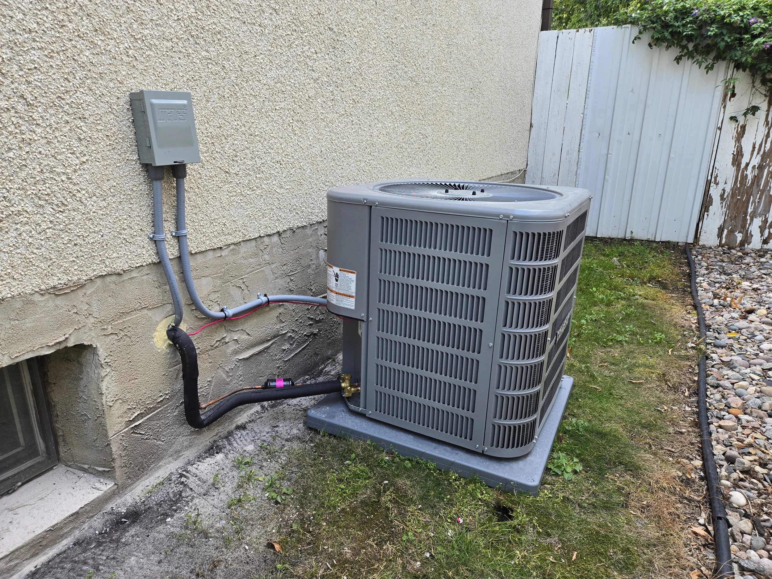 Outdoor air conditioning unit next to a textured stucco wall, with electrical connections and a small black pipe, on a concrete slab with a grassy area and a white wooden fence nearby.