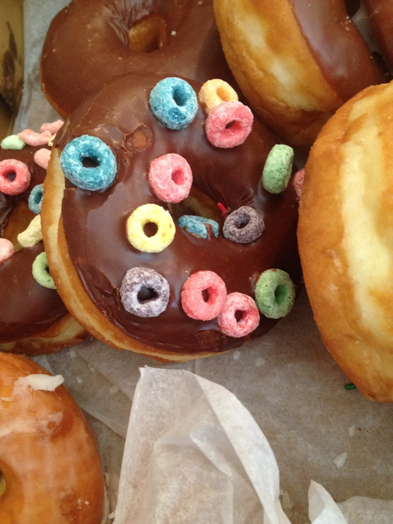Chocolate-glazed donuts topped with colorful cereal pieces, placed on parchment paper.