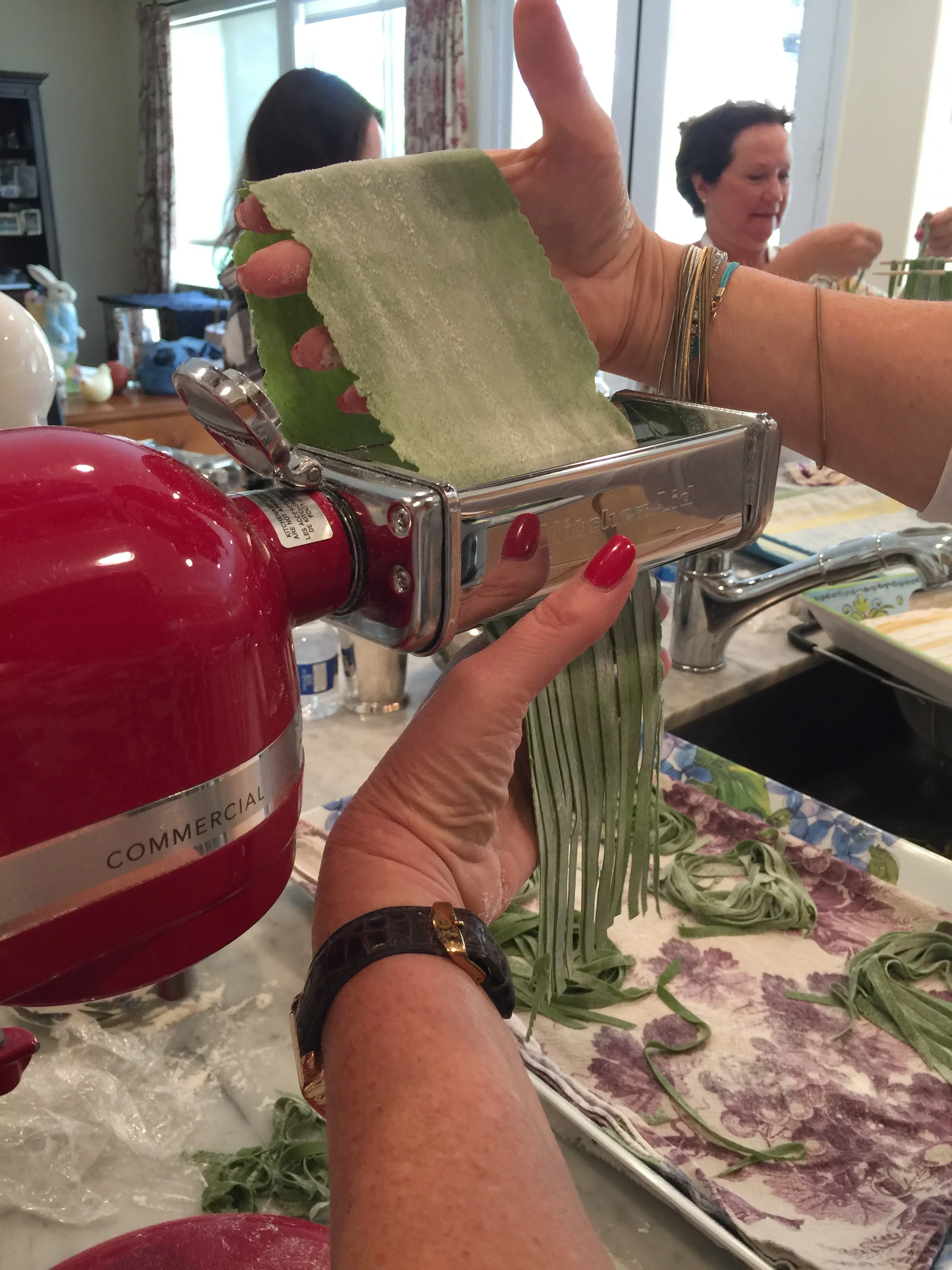 A person pressing fresh green handmade pasta through a pasta machine, with other people in the background in a kitchen.