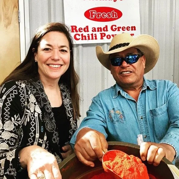 A woman and a man are smiling at the camera at a chili tasting event, with a sign behind them that reads 'Fresh Red and Green Chili Powder.' The man is wearing a cowboy hat, sunglasses, and a denim shirt, and is holding a bowl of red chili powder.