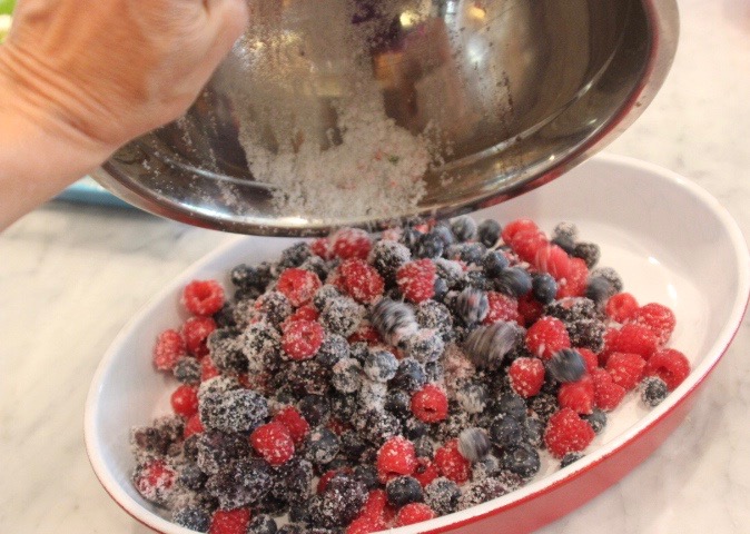 Frosted mixed berries, including raspberries and blueberries, in a white oval dish, with a person sprinkling powdered sugar from a small metal bowl.