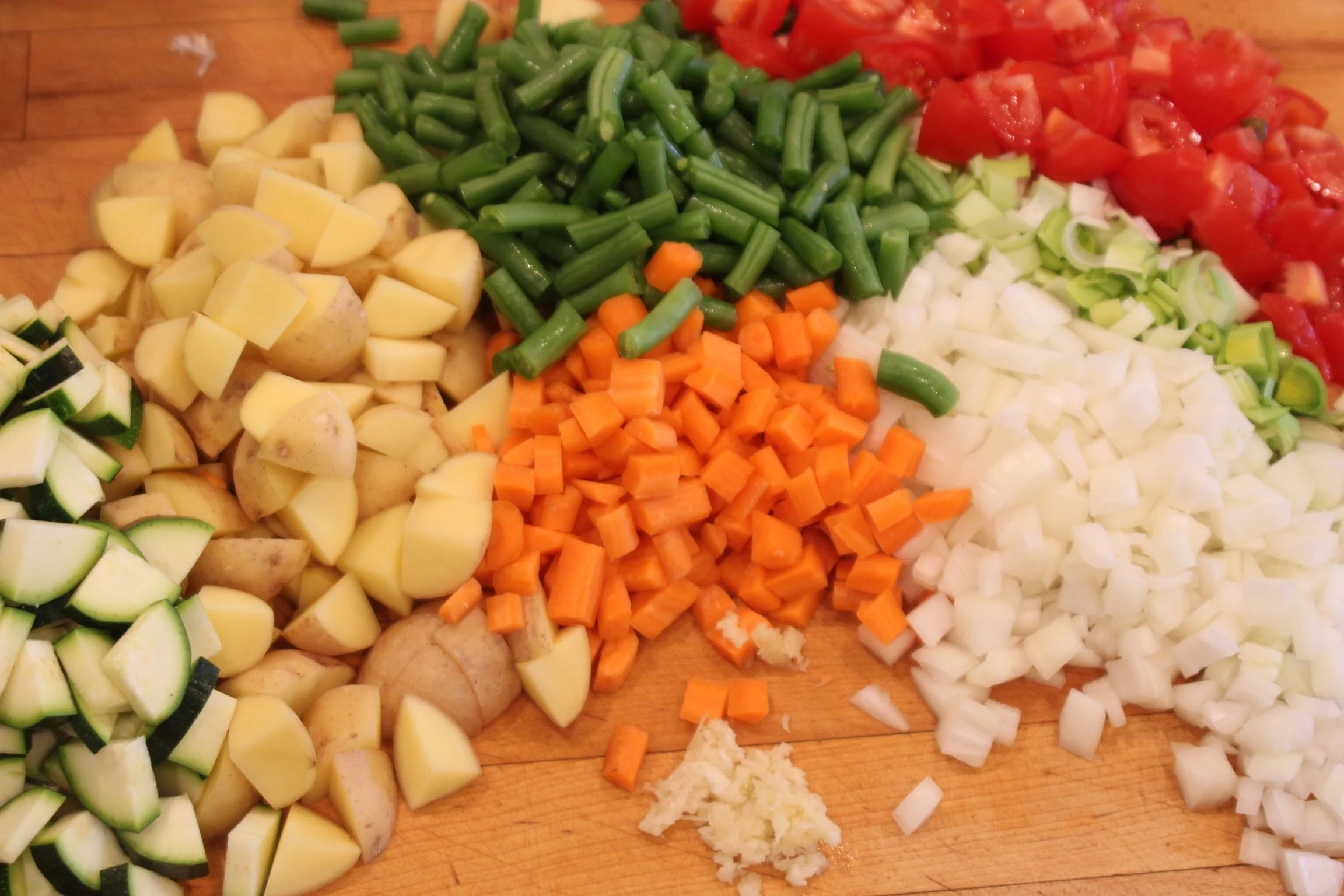 Chopped vegetables including zucchini, potatoes, carrots, onions, tomatoes, and celery on a wooden cutting board.