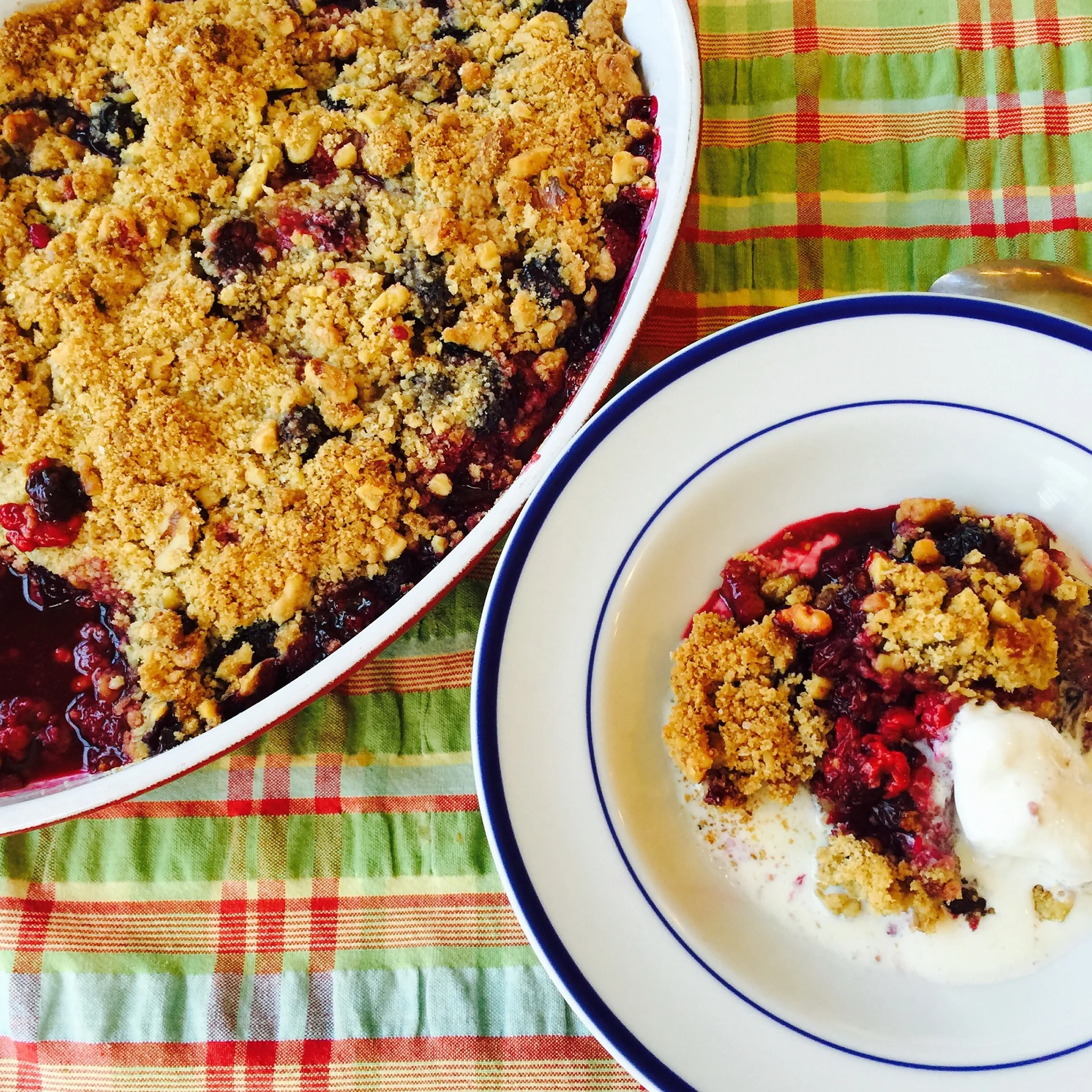 A cherry or berry cobbler in a rectangular baking dish with a crumbly topping, served with a bowl of vanilla ice cream on a green and red plaid tablecloth.