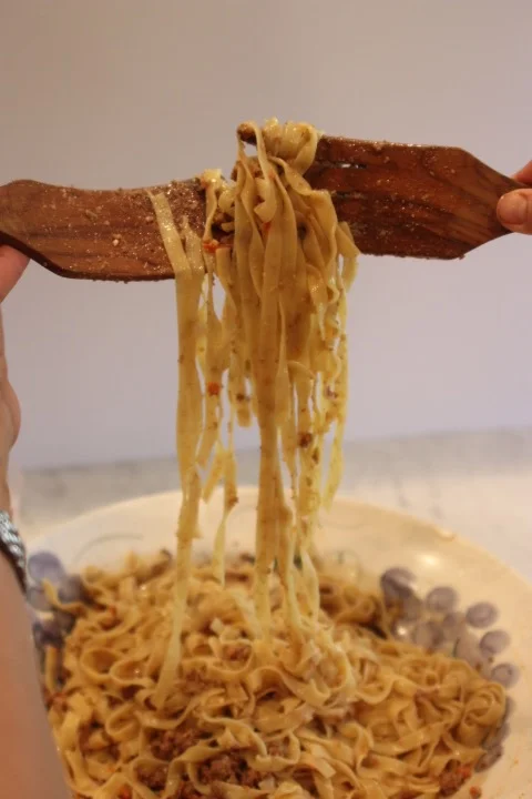 Person holding a large piece of wood with cooked noodles hanging from it, over a plate of cooked pasta and peas.
